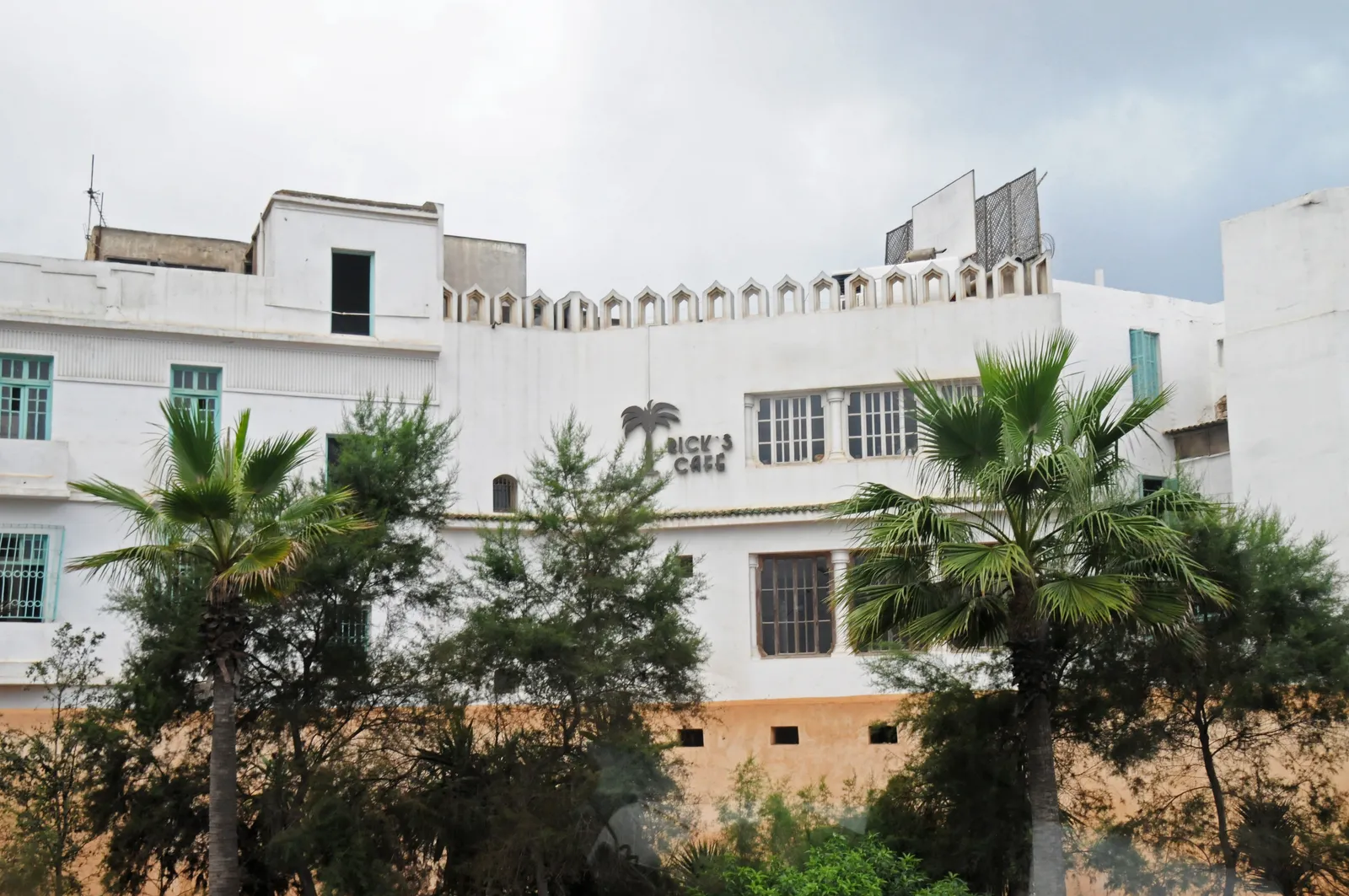 The white walls and crenellated roofline of Rick's Café in Casablanca seen over a screen of palms, with the name lettered on the upper wall beside a row of arched openings