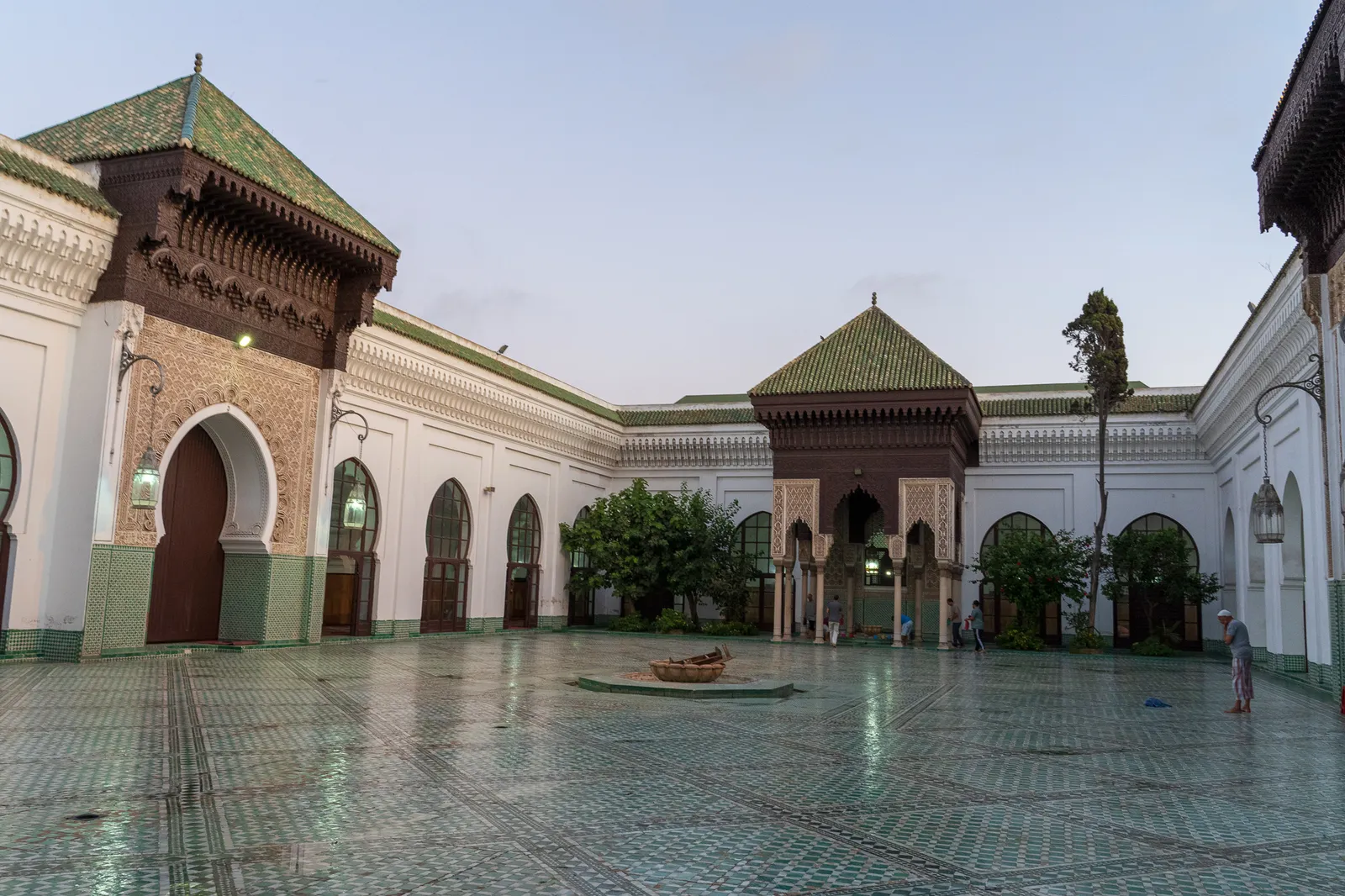 The courtyard of the Al-Mohammadi Mosque in Casablanca's Habous quarter, green tiled roofs over carved cedar eaves, horseshoe-arched gateways on two sides and a patterned tile floor running the width of the court