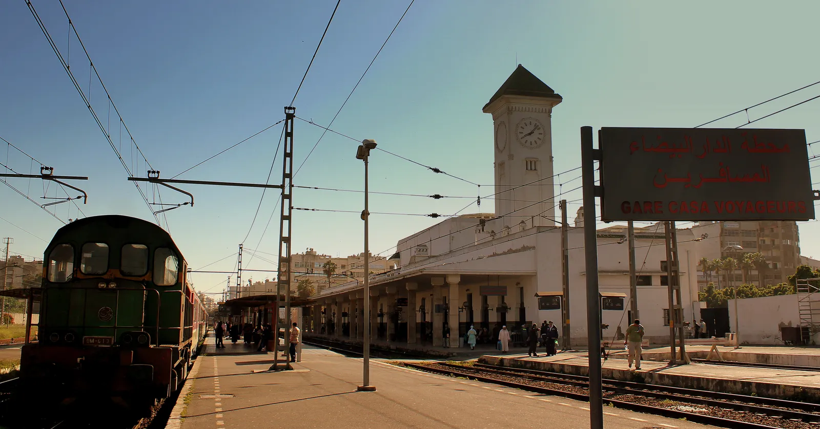 The 1923 station building at Casa-Voyageurs seen from the platforms, a white hall with arched windows under a square clock tower, overhead wires above the tracks and a green locomotive standing at the left
