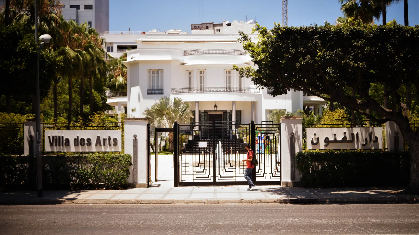 The Villa des Arts in Casablanca seen from the street, a white 1930s villa with a rounded corner and deep balconies standing behind iron railings, its name lettered in Latin and Arabic script along the garden wall