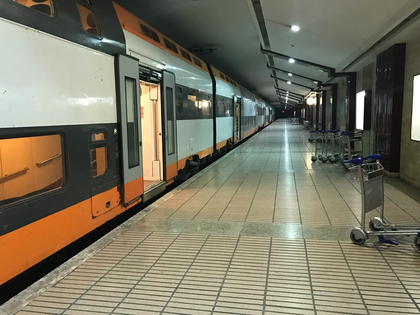 The underground railway platform at Mohammed V International Airport, an orange and white double-deck train standing at the edge and a row of luggage trolleys lined up along the tiled floor