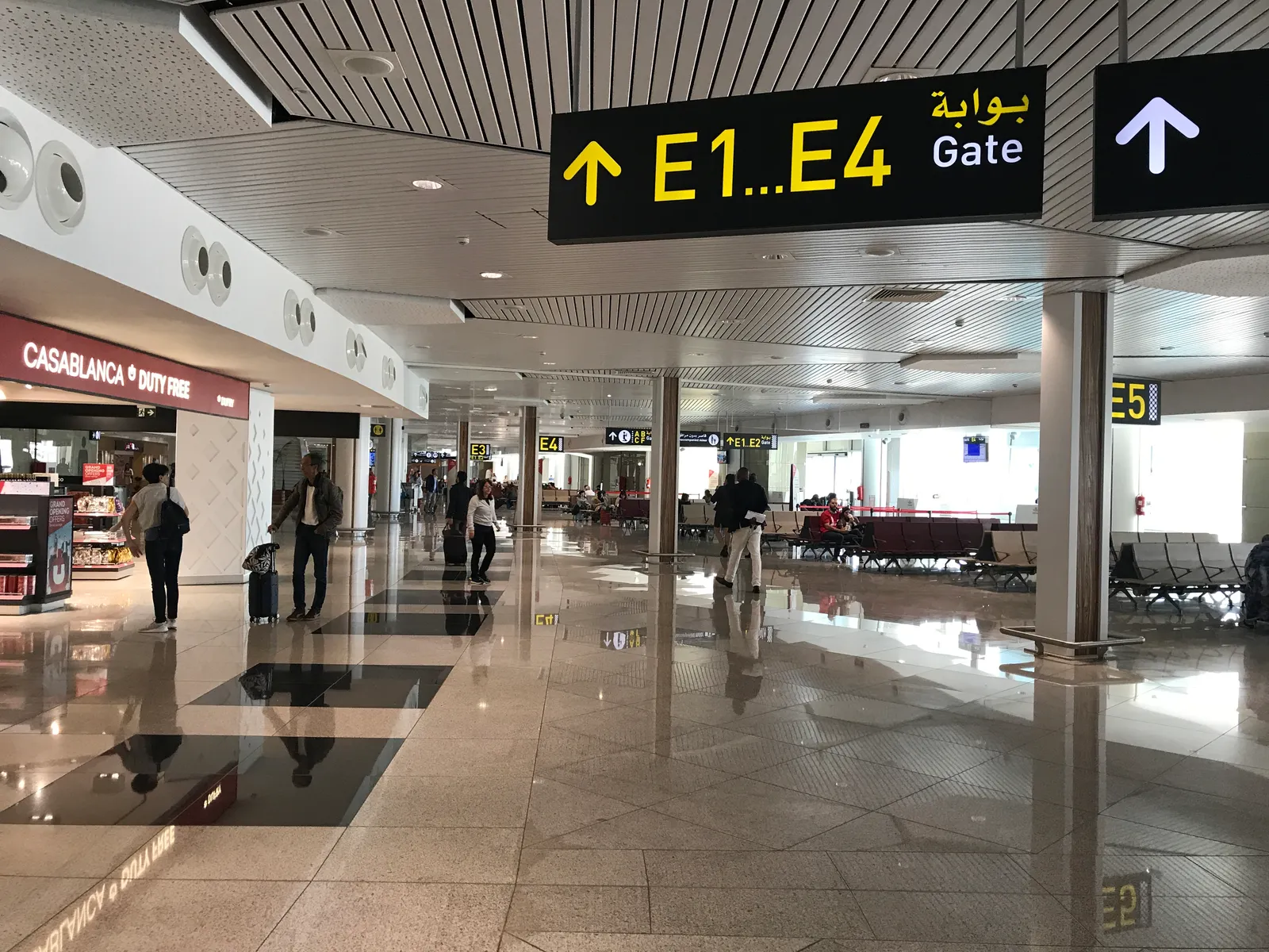 The departures concourse in Terminal 1 at Mohammed V International Airport, an overhead sign pointing to gates E1 to E4 in Arabic and English, a duty-free shop on the left and rows of seating beyond