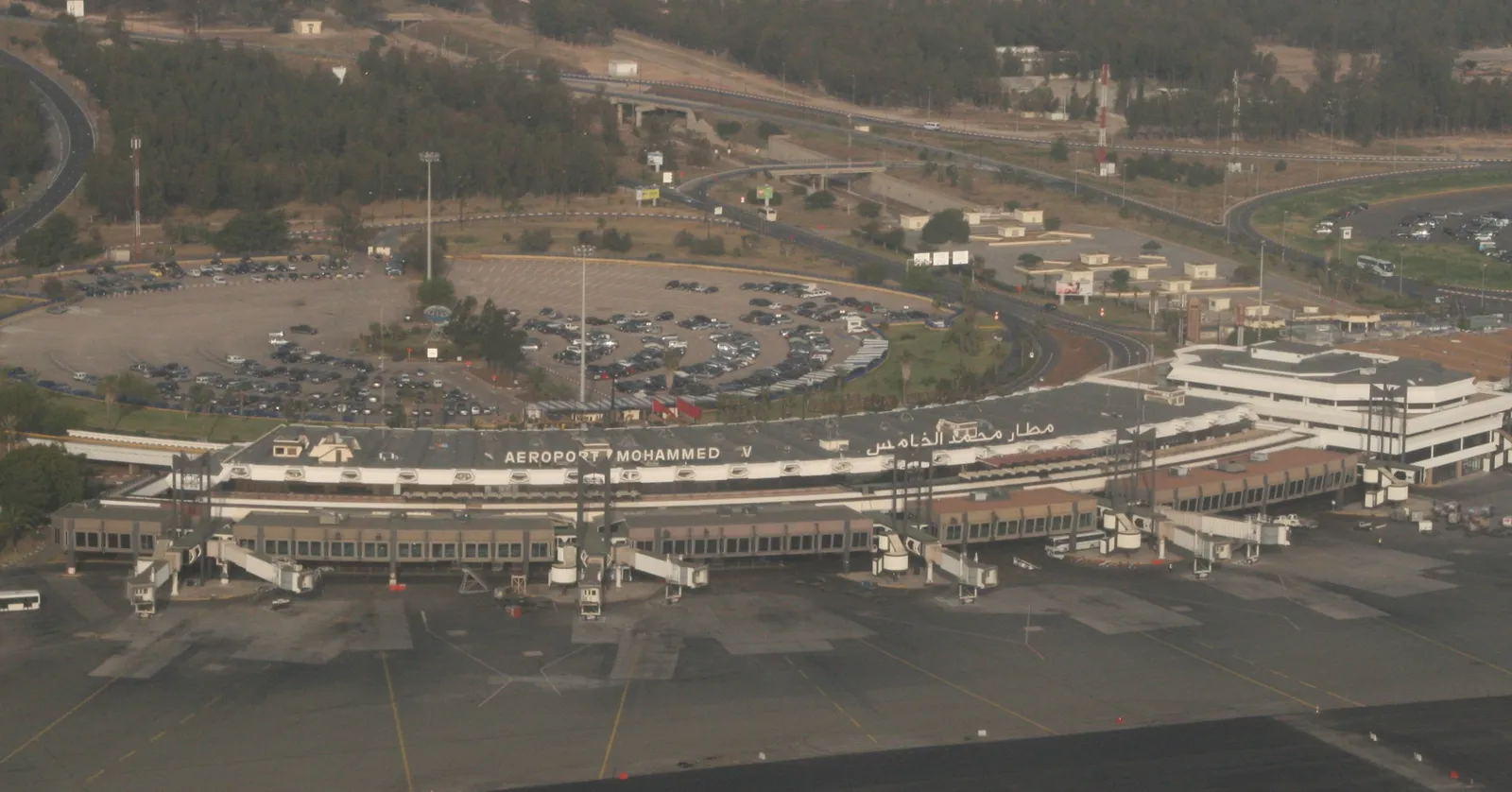 Mohammed V International Airport seen from the air, the long low terminal building with AEROPORT MOHAMMED lettering across its front, jet bridges reaching out to the apron and car parks and motorway behind it