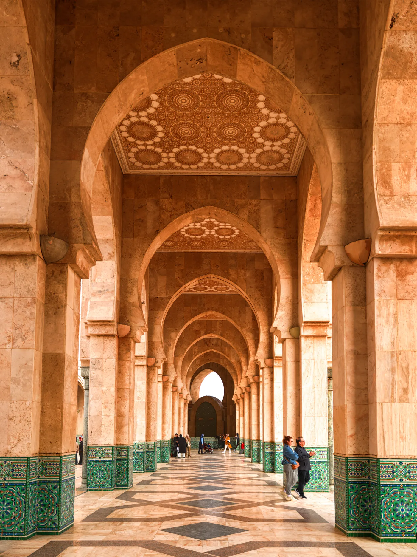 An arcade at the Hassan II Mosque, a long run of horseshoe arches on sandstone piers receding into the distance, carved plasterwork in the vaults, green and white zellige along the base of the walls and a star-patterned marble floor