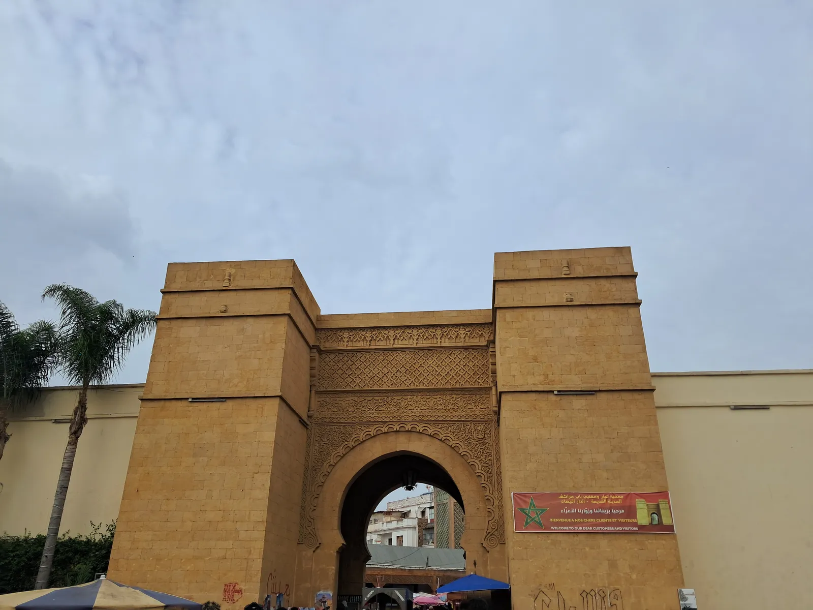 Bab Marrakech, the sand-coloured southern gate of Casablanca's old medina, a tall horseshoe arch carved with geometric bands between two square towers, with market awnings at its foot