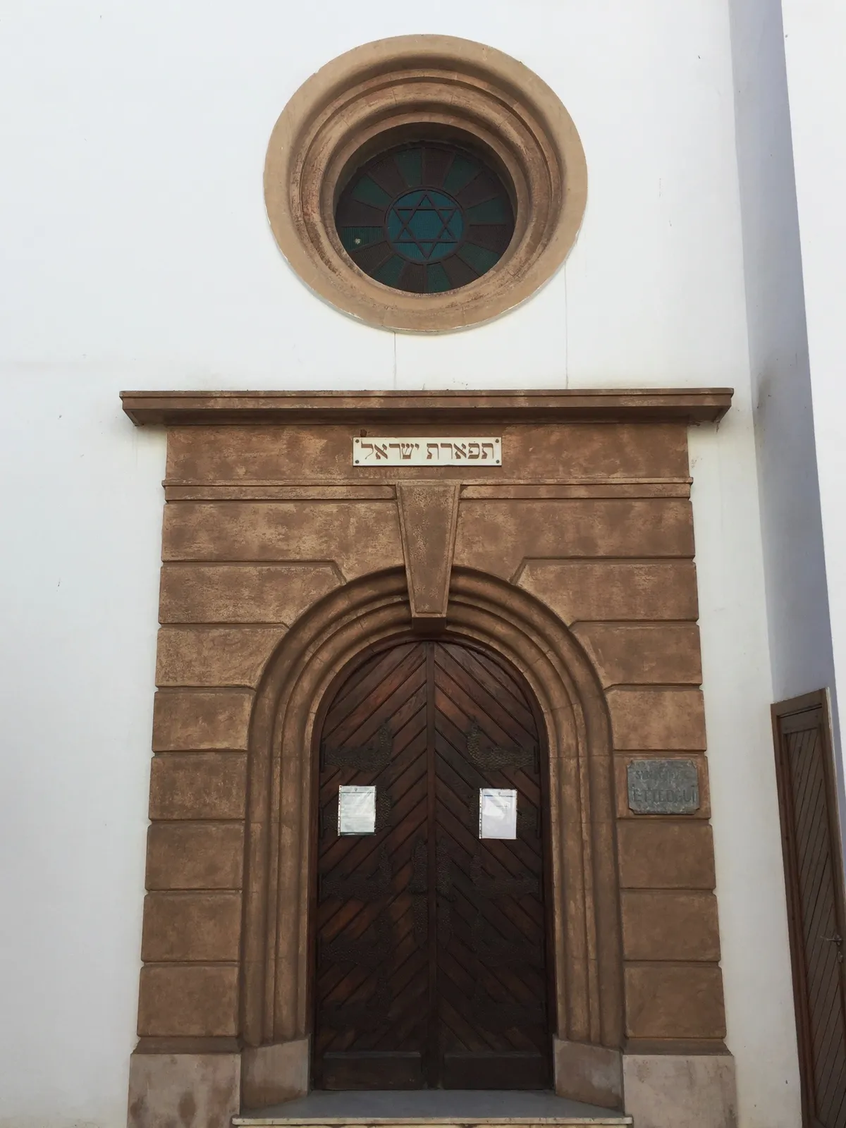 The entrance to the Ettedgui Synagogue in Casablanca, a stone arched doorway with heavy wooden doors, a Hebrew inscription on the lintel above it and a round window holding a stained-glass Star of David set into the white wall