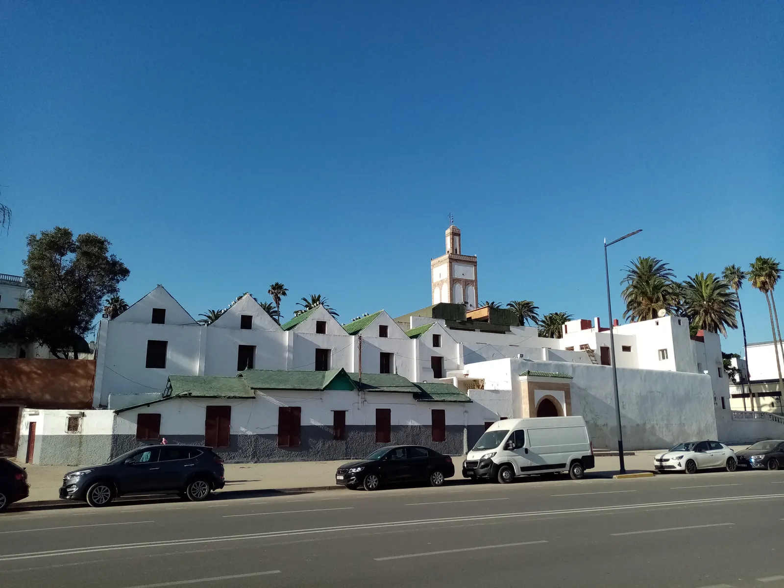 The Ould el-Hamra Mosque in Casablanca's old medina seen from the street, whitewashed walls under pitched green-tiled roofs with an arched doorway, and a square minaret with green tilework standing behind