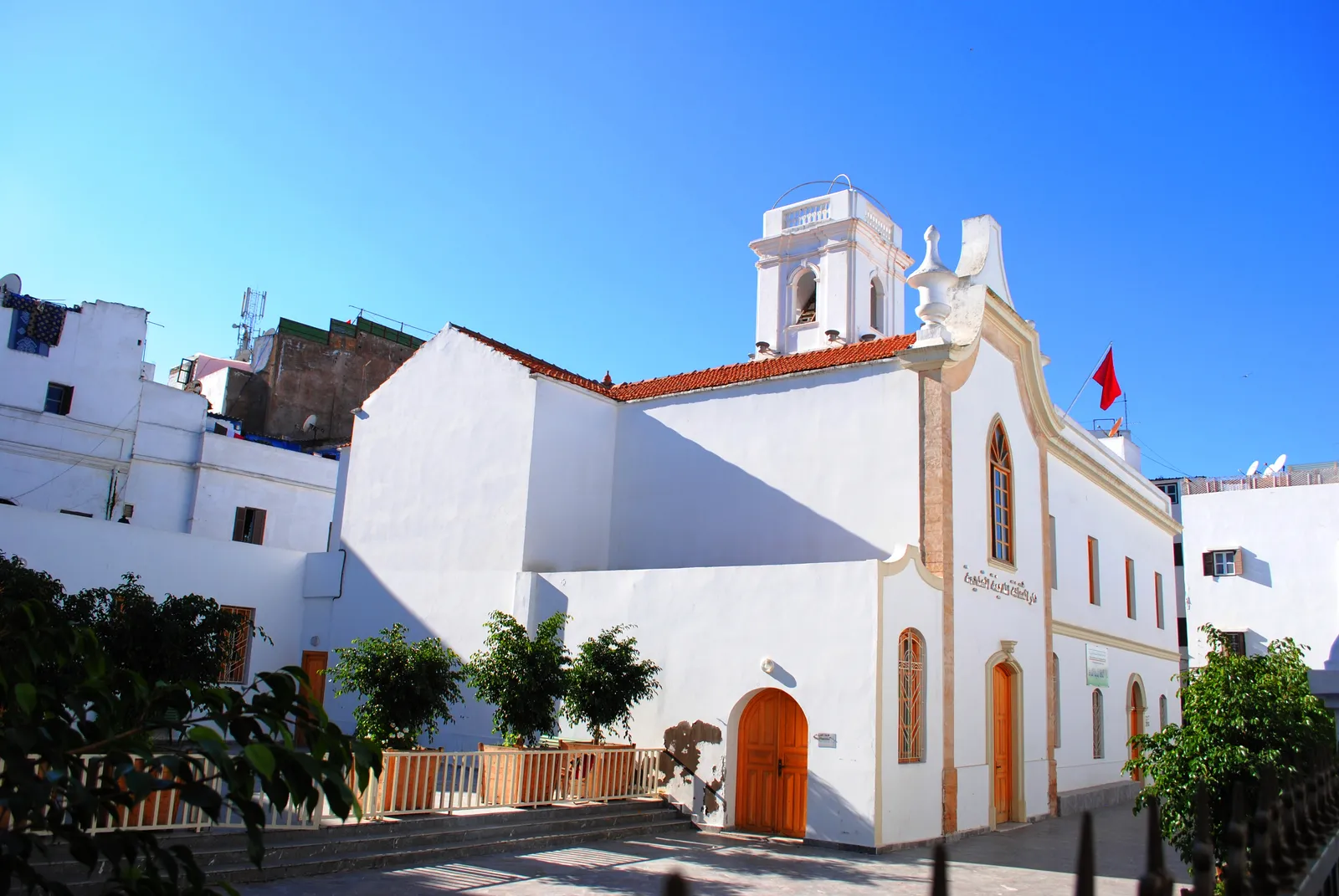 The Church of San Buenaventura in Casablanca's old medina, a whitewashed building with a red-tiled roof, a small white bell tower and pointed arched windows, standing above a fenced garden in a narrow square