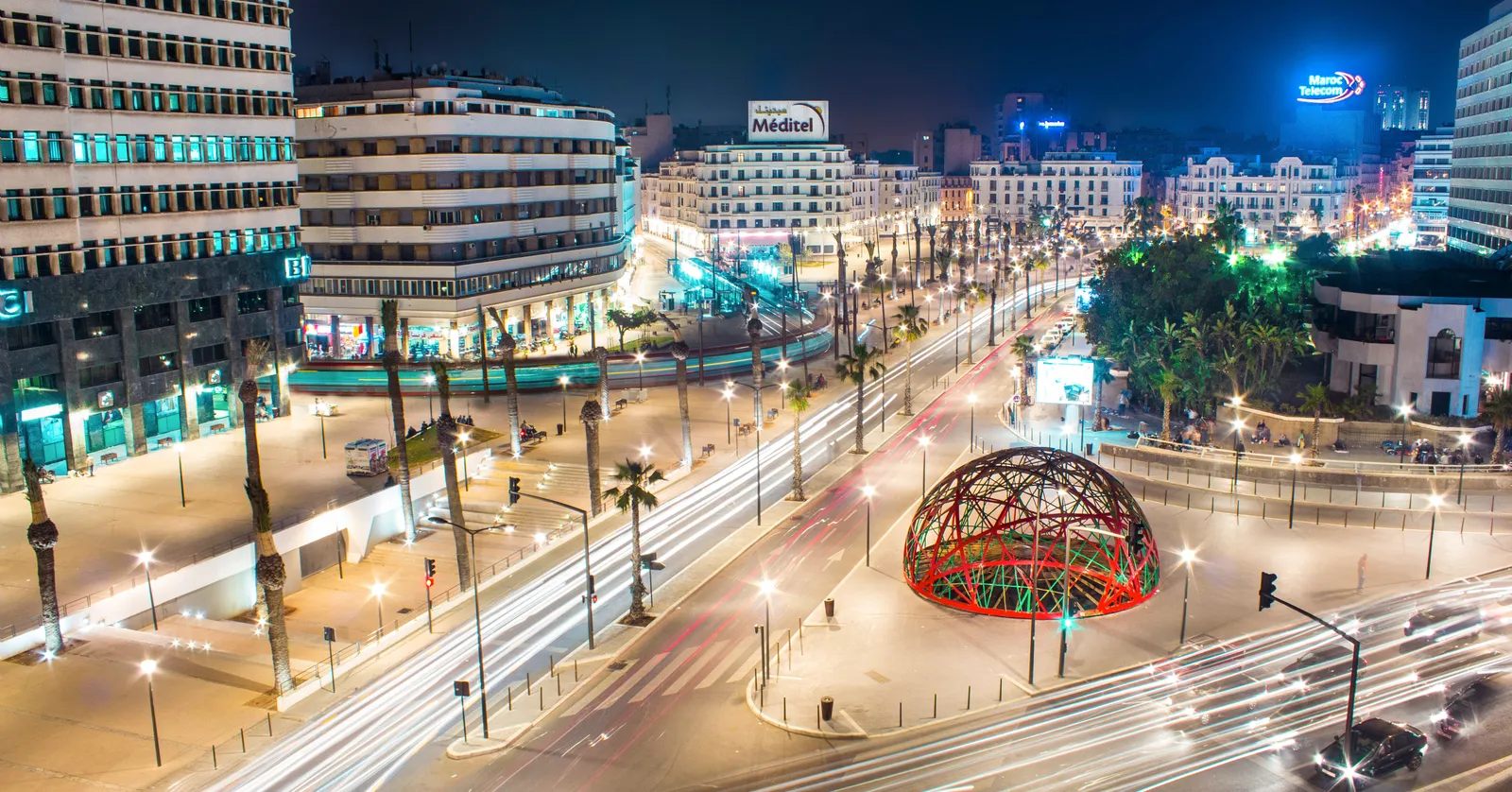 United Nations Square in Casablanca at night seen from above, a red steel lattice half-globe lit up in the middle of the paving, tram tracks and light trails from traffic curving around it and 1930s apartment blocks along the edges
