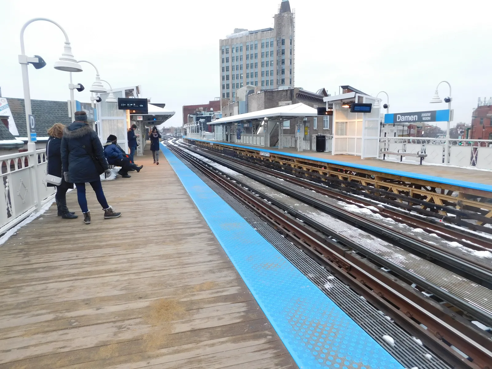 The wooden side platform at Damen station in Chicago under snow, with the blue platform edge strip, gooseneck lamps along it and the Art Deco tower at the six corners in the background
