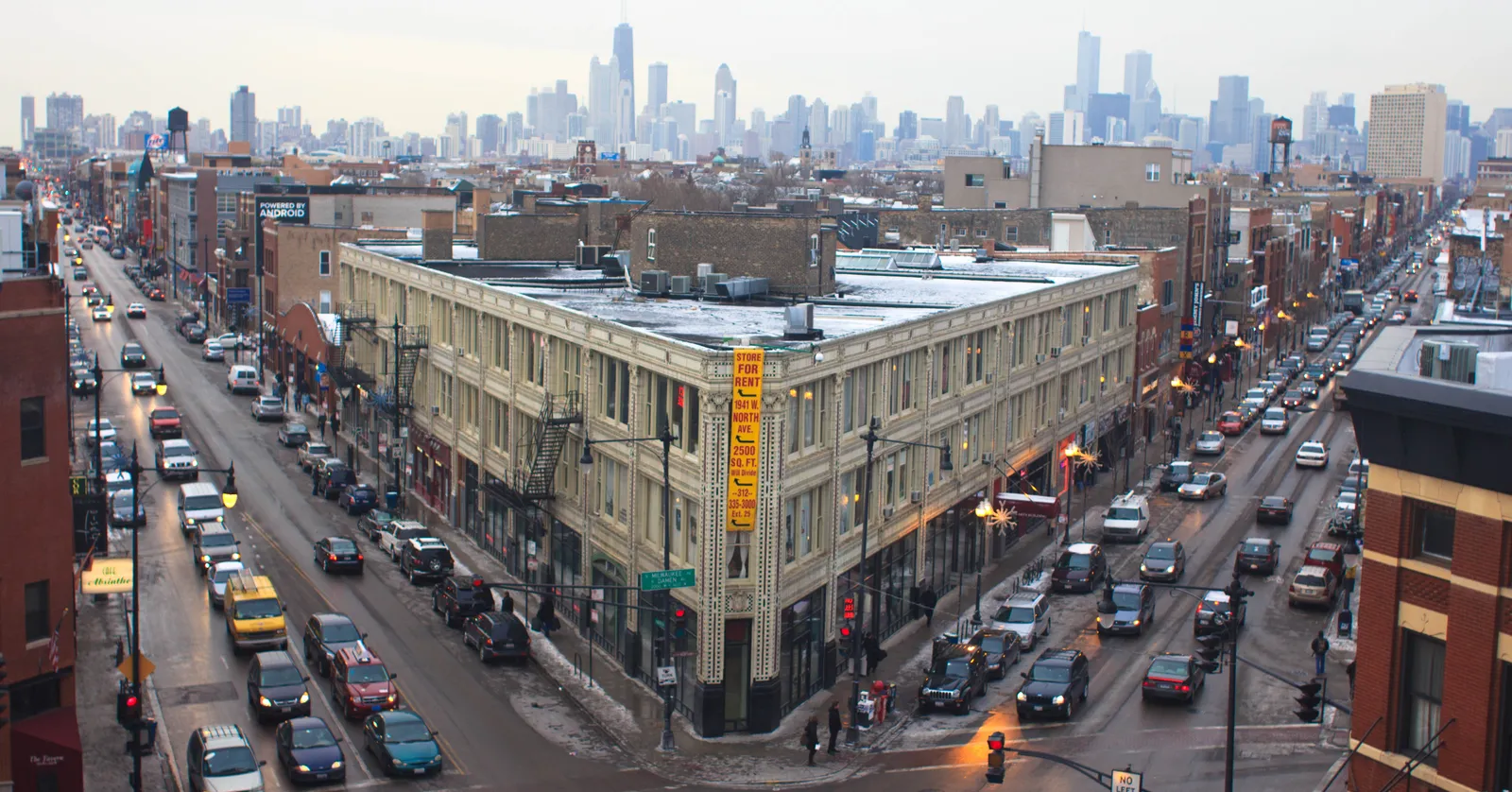The cream terracotta Flat Iron Arts Building on its triangular corner in Chicago's Wicker Park, seen from above with traffic on Milwaukee Avenue either side of it and the Loop skyline on the horizon