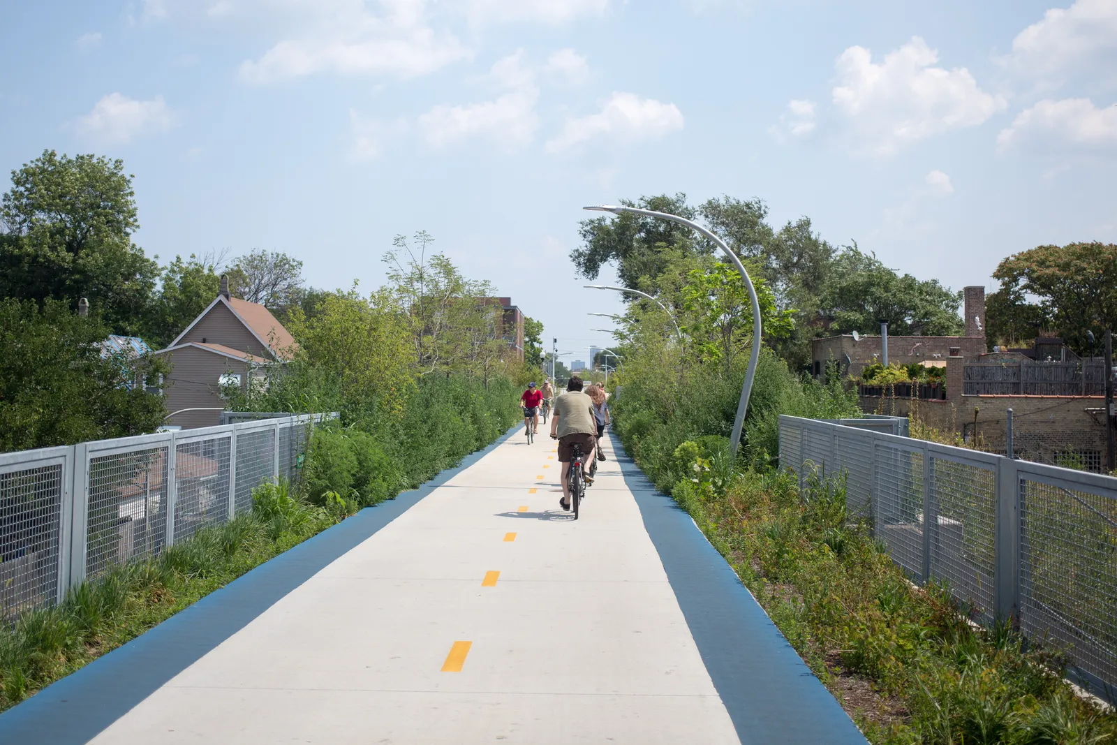 The concrete path of the 606 trail in Chicago running straight ahead between planted borders and steel fences, a cyclist and two walkers on it and curved lamp posts arching overhead