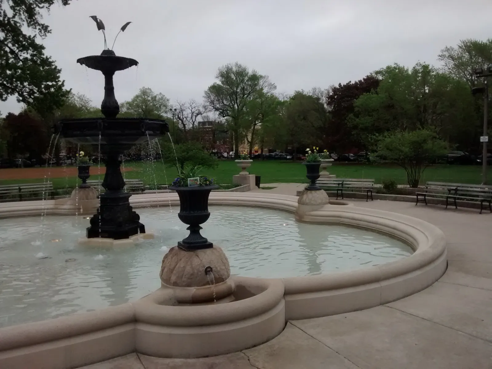 The tiered black iron fountain in Wicker Park in Chicago, standing in a pale stone basin with planted urns around the rim, benches and lawn behind it