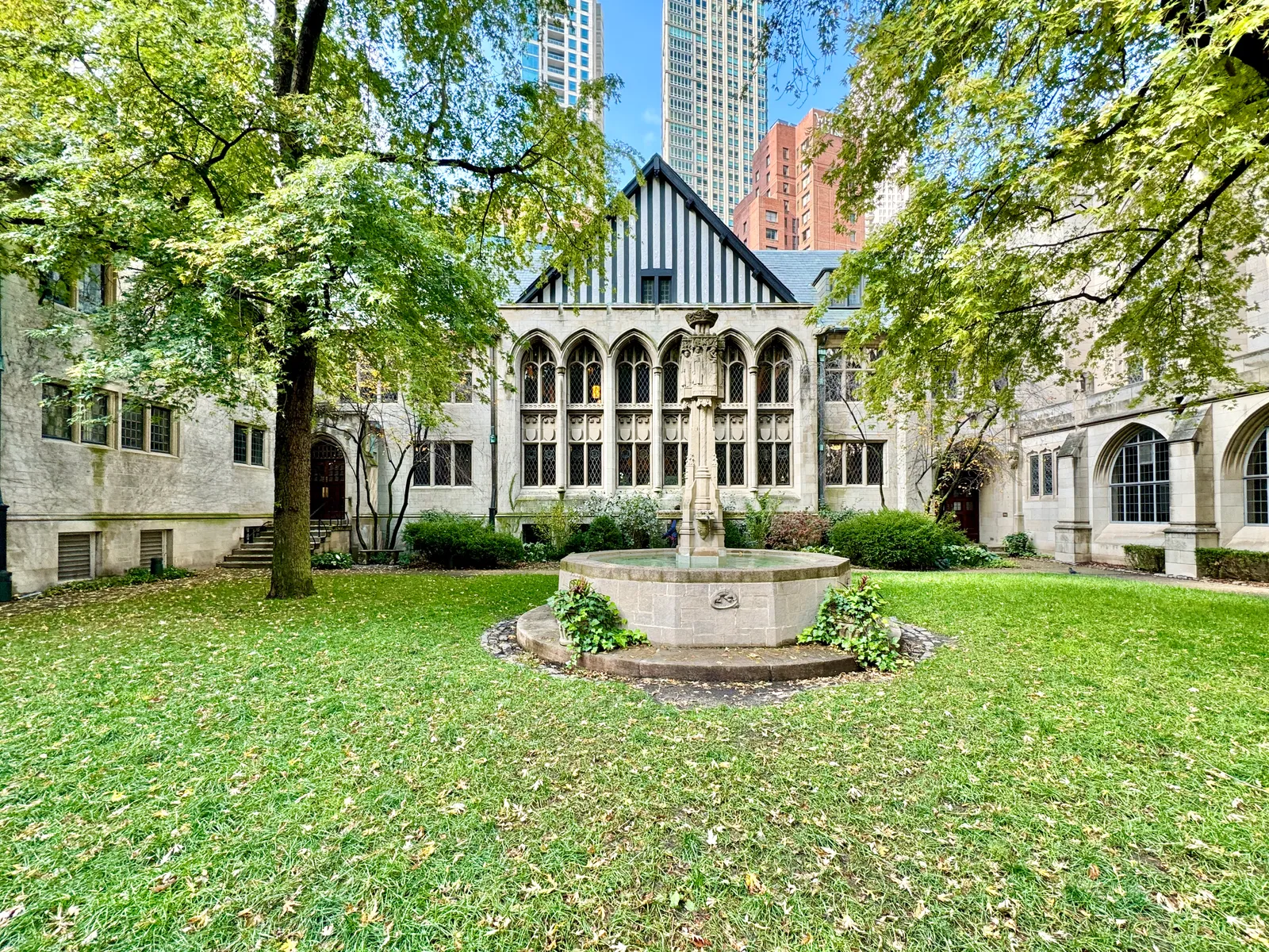 The walled cloister garden at Fourth Presbyterian Church in Chicago, a lawn with a stone fountain in the middle, Gothic arcades on two sides and high-rise towers visible over the roofline