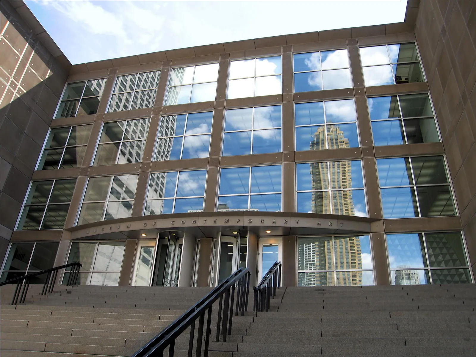 The glass and limestone front of the Museum of Contemporary Art in Chicago, with a broad flight of stone steps leading up to its entrance and the neighboring towers reflected in the windows
