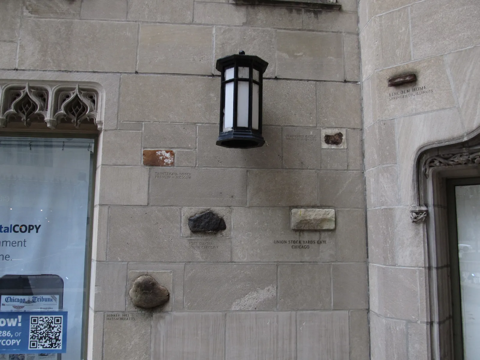 Fragments of stone from famous buildings set into the limestone wall at the base of Tribune Tower in Chicago, each one labeled with a small engraved caption, below an iron lantern