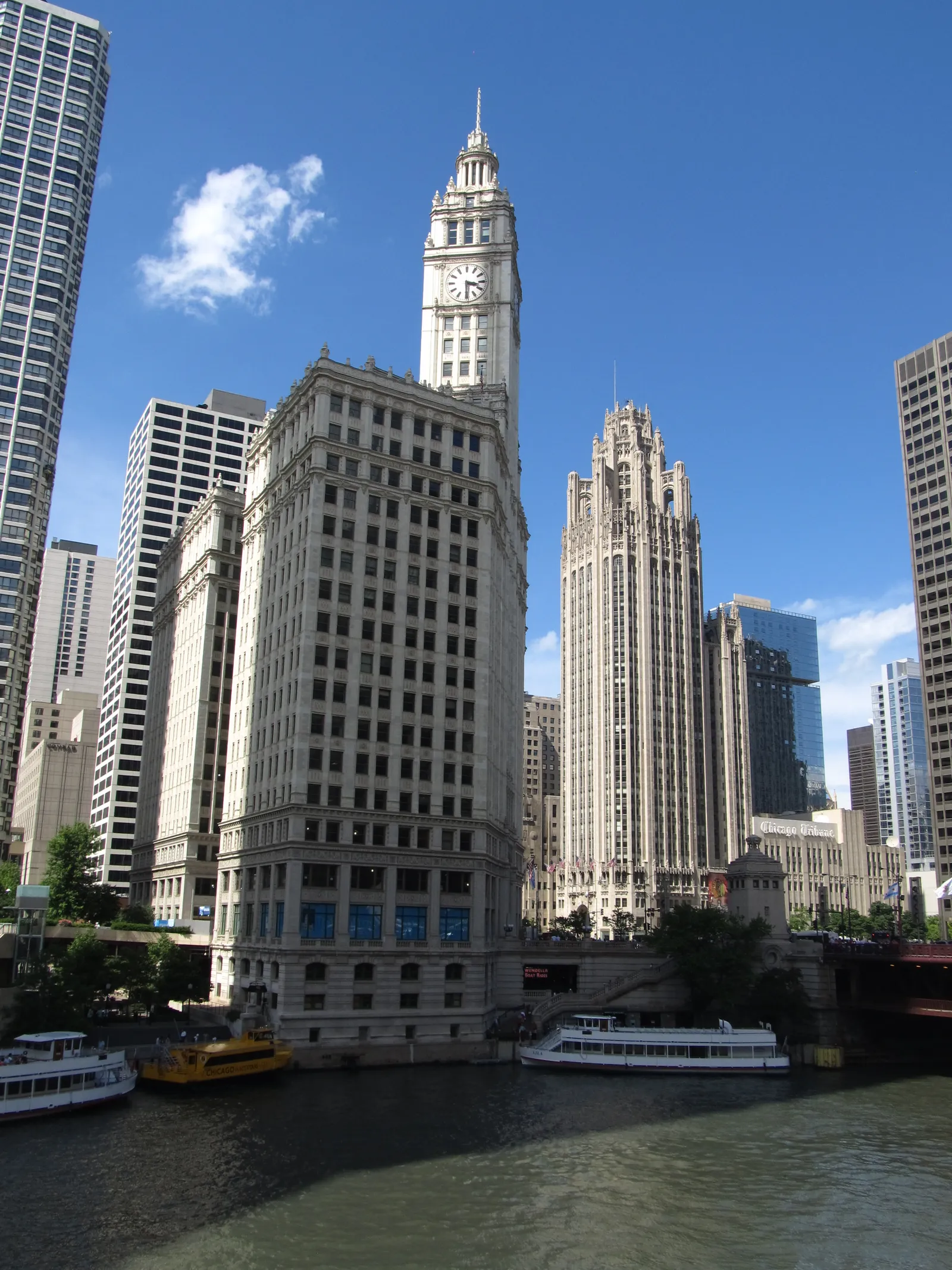 The white terracotta Wrigley Building and its clock tower on the north bank of the Chicago River, with Tribune Tower's stone crown beside it and sightseeing boats moored at the bridge below