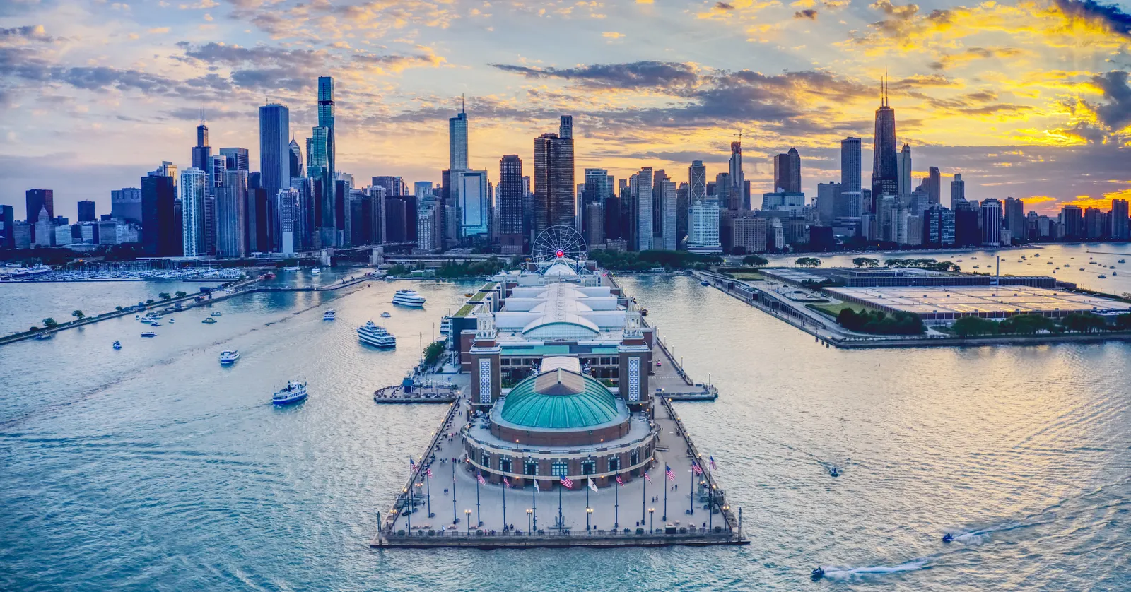 Navy Pier seen from above the lake at sunset, its long deck running back toward the Chicago skyline, with the domed ballroom at the near end and the Ferris wheel behind it