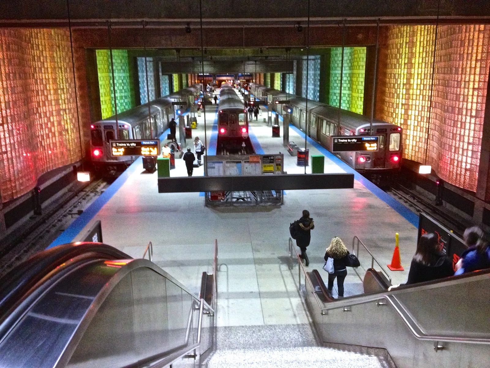 Blue Line trains waiting at the O'Hare station platform in Chicago, between walls of backlit glass block in shifting colors, seen from the top of the escalator