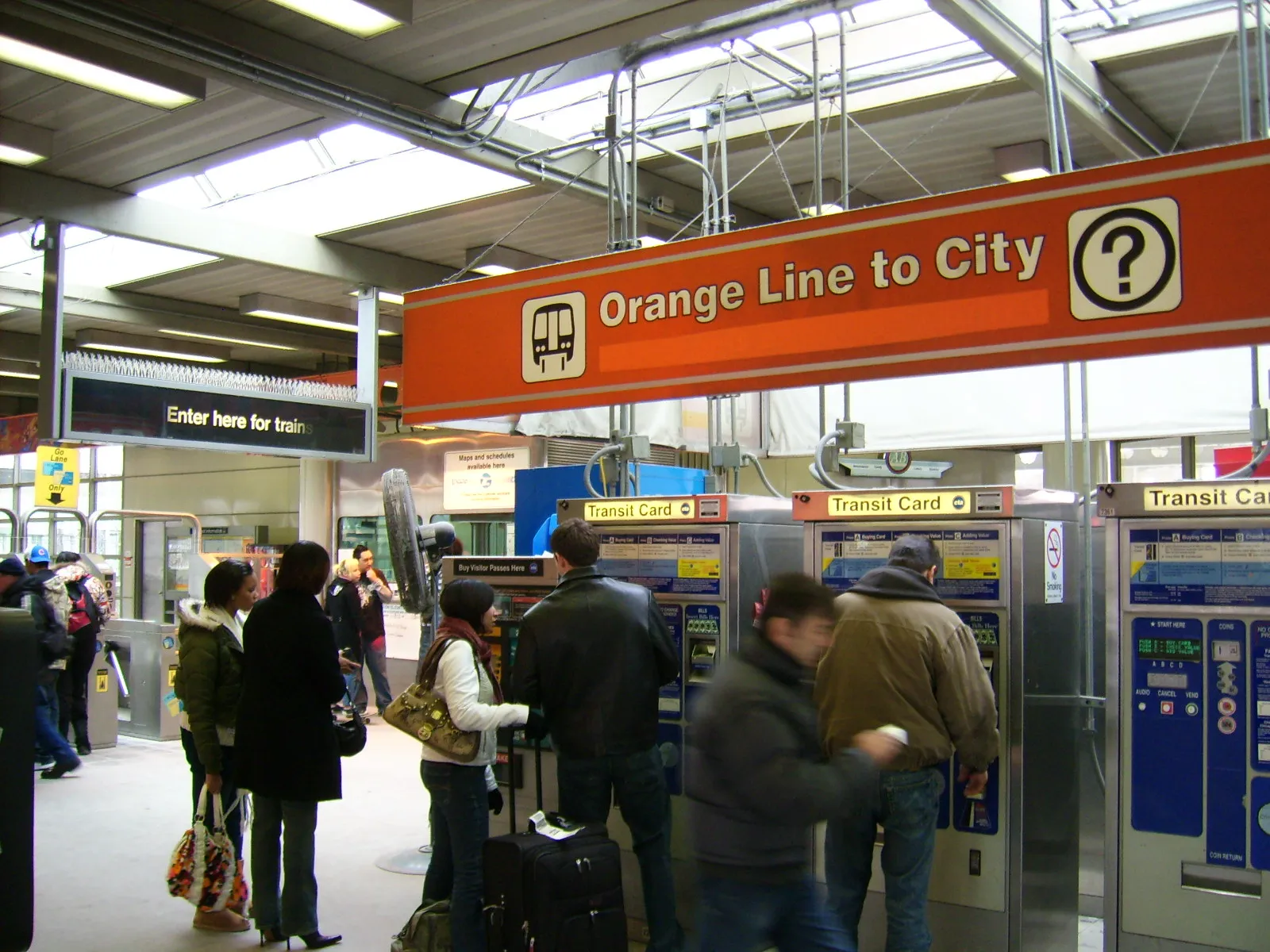 Passengers with suitcases at the ticket machines of the Midway Orange Line station in Chicago, under an orange sign reading Orange Line to City