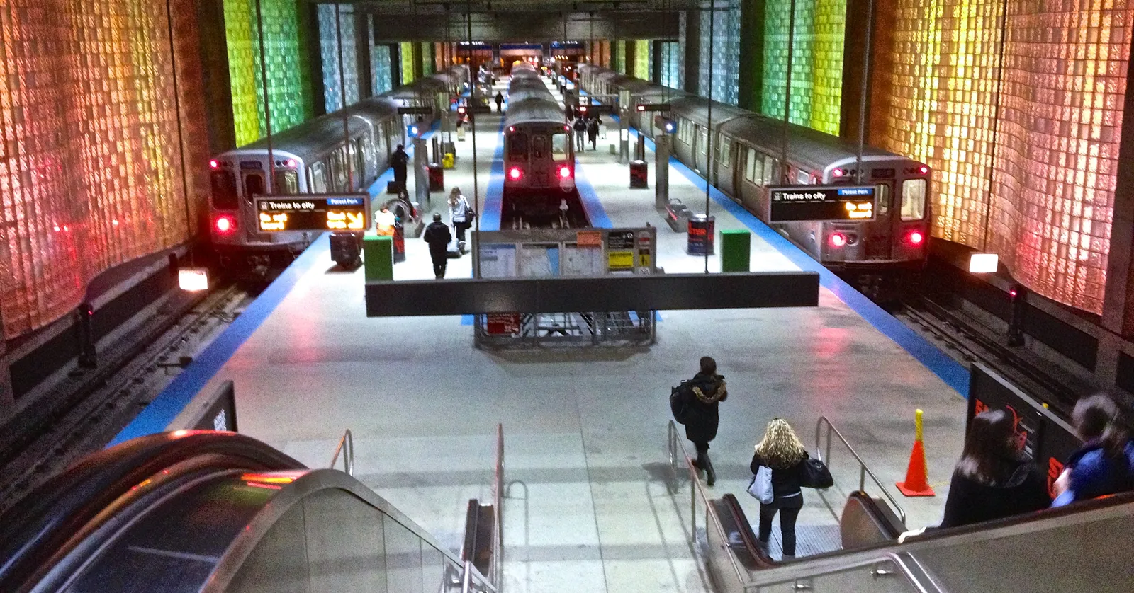 Blue Line trains waiting at the underground O'Hare station in Chicago, between walls of backlit coloured glass block, seen from the top of the escalator