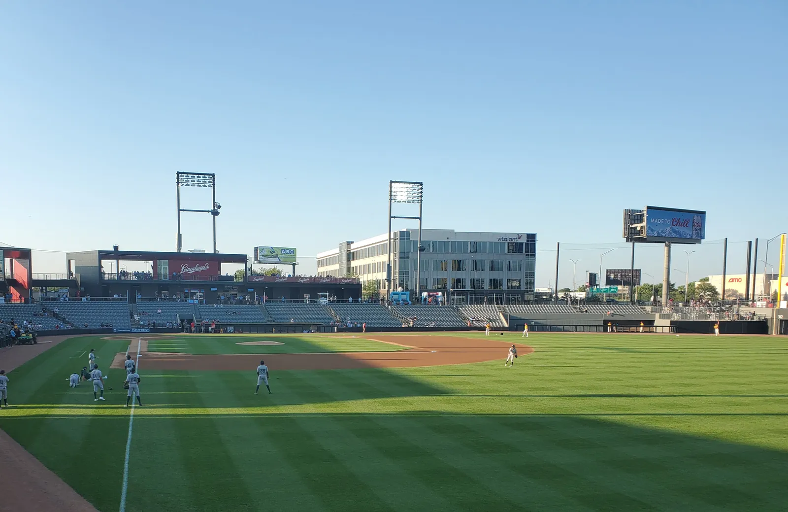 A baseball game under way at Impact Field in Rosemont, seen from the outfield grass, with the grandstand, floodlight masts and a video scoreboard behind the infield