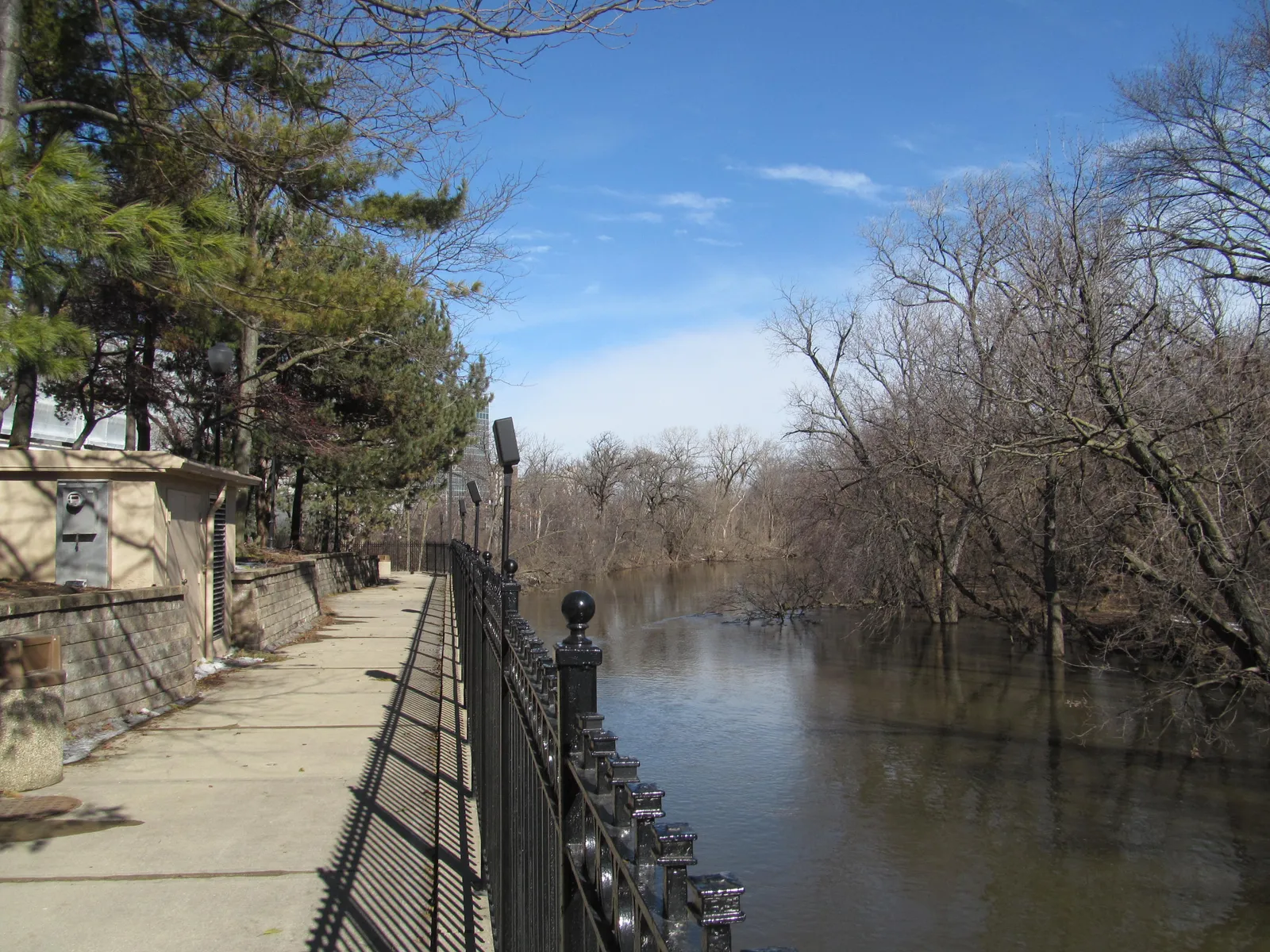 A paved walkway with a black iron railing running along the wooded bank of the Des Plaines River at Monument Park in Rosemont, bare trees on both sides of the water
