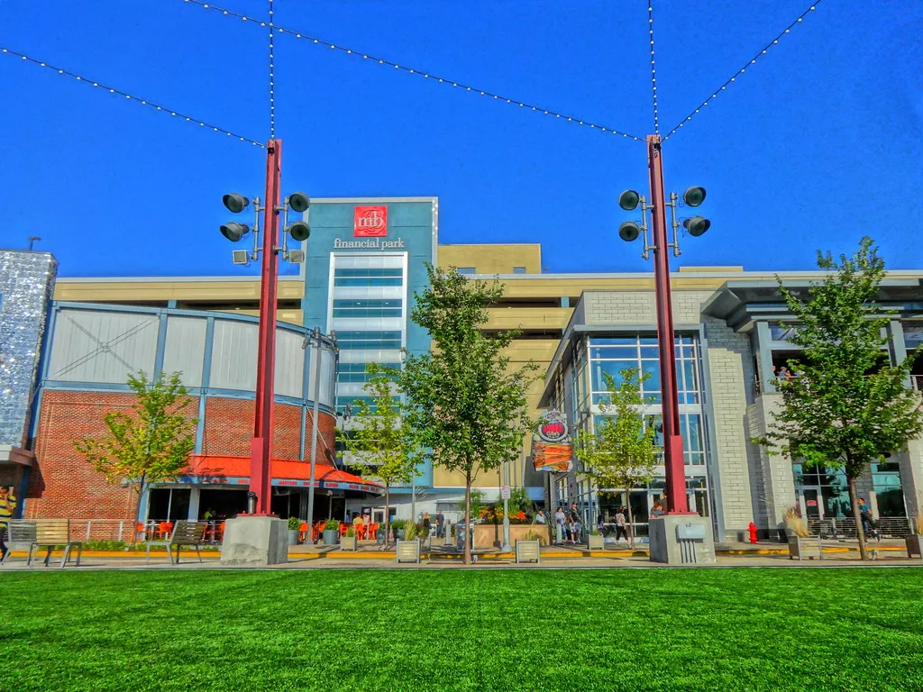 The open lawn at Rosemont's entertainment district, with bar and restaurant frontages along the far side, tall red floodlight masts and strings of lights crossing a blue sky
