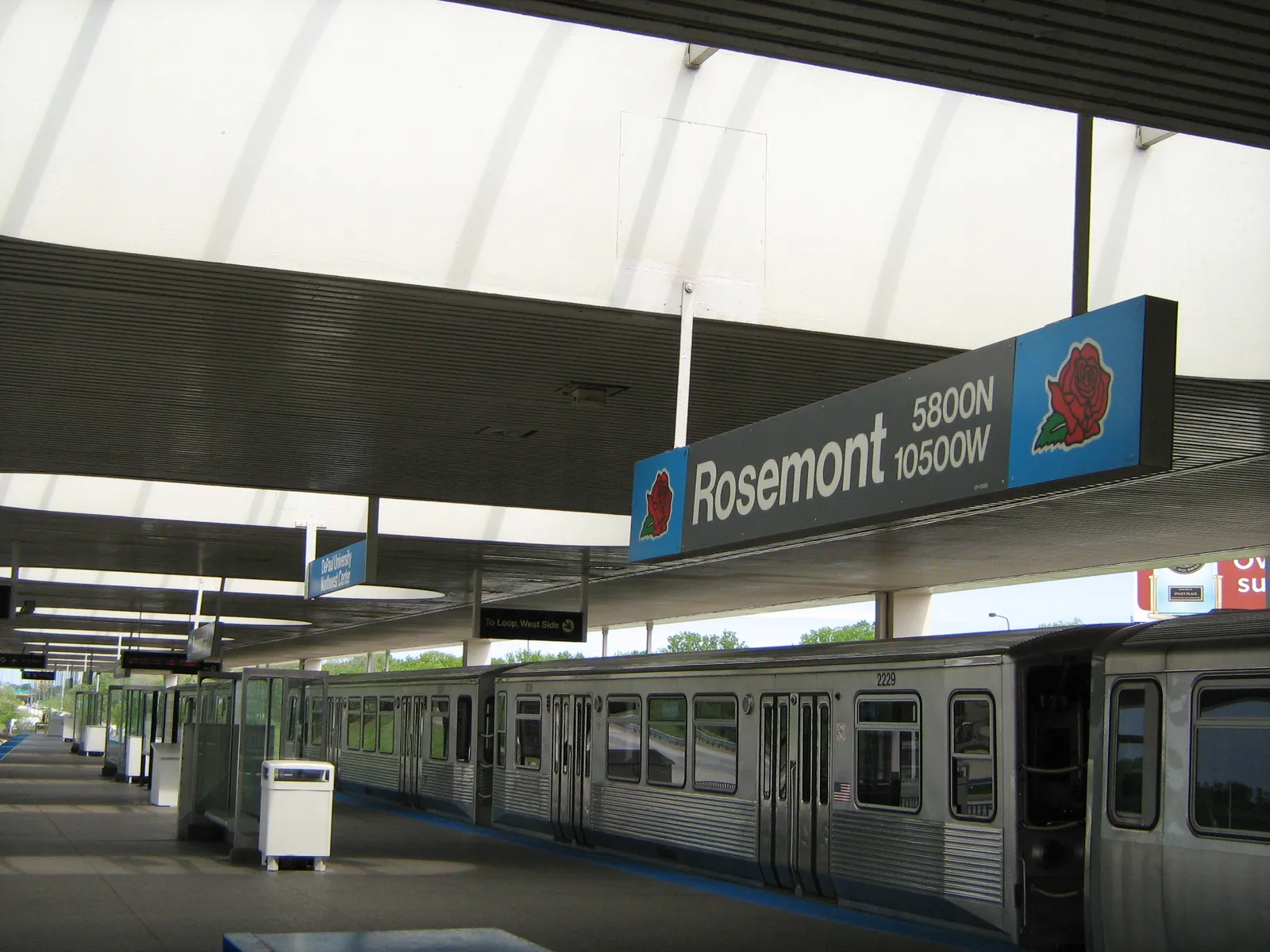 The island platform at Rosemont station on Chicago's Blue Line, a train standing at the edge and a hanging sign carrying the station name beside the village's red rose emblem