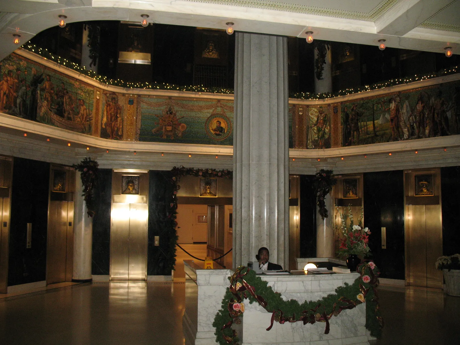 The two-story lobby of the Marquette Building in Chicago, with a band of Tiffany glass mosaic panels running around the balcony rail above bronze elevator doors and dark marble walls