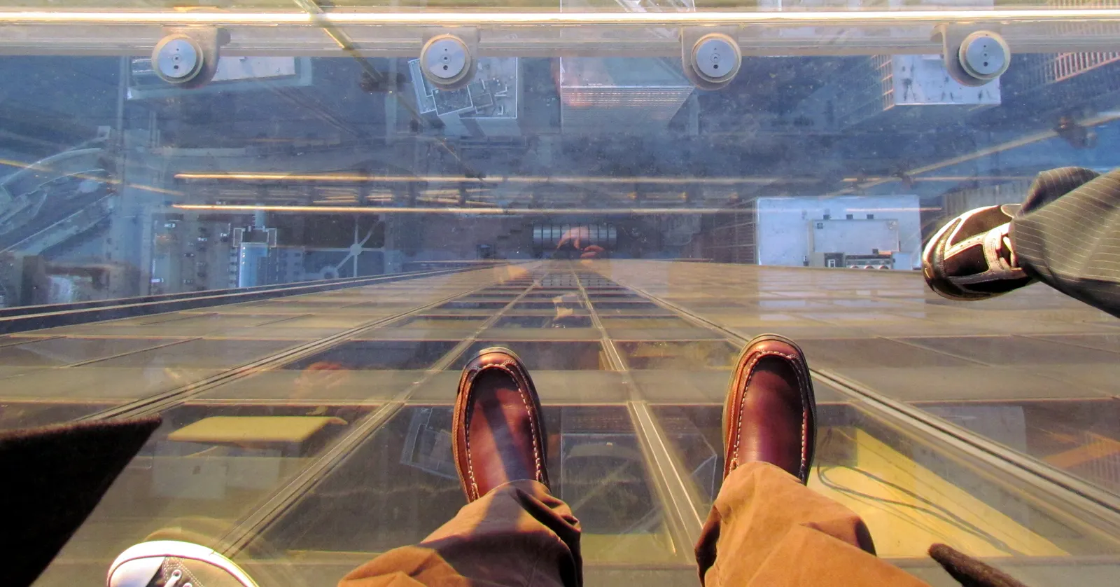 Looking straight down through the glass floor of a Skydeck ledge box at Willis Tower, with two visitors' feet on the glass and Chicago streets and rooftops far below