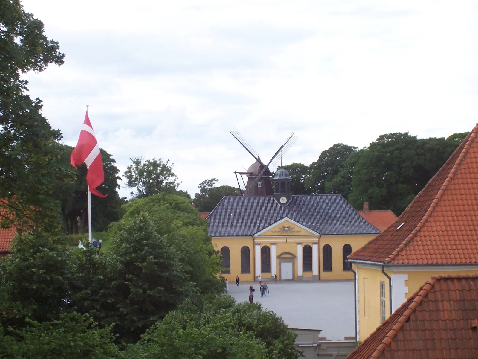 Inside Kastellet in Copenhagen, a yellow church with a small spire standing on the parade ground and a black wooden windmill on the grass rampart behind it, with a Danish flag flying in the foreground