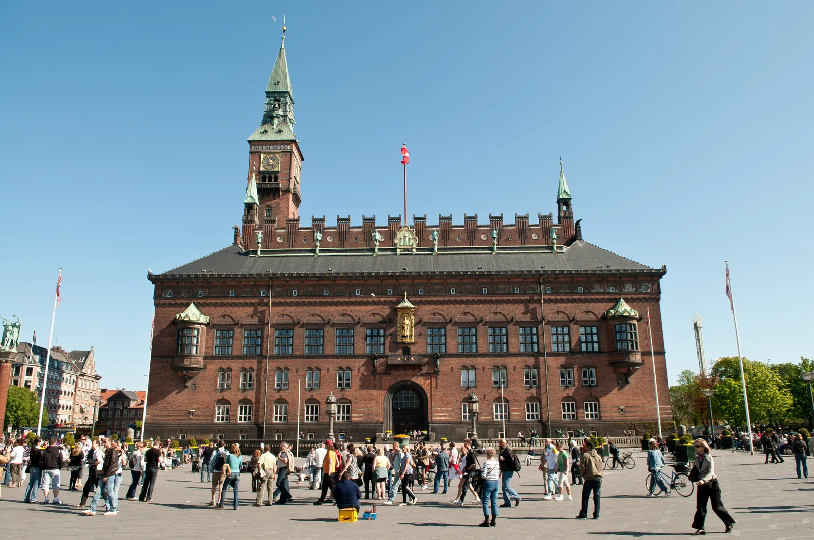 Copenhagen City Hall seen across its open square on a sunny day, a long red brick front with a gilded figure above the entrance and a green-spired clock tower rising at the left end, with people crossing the paving in front
