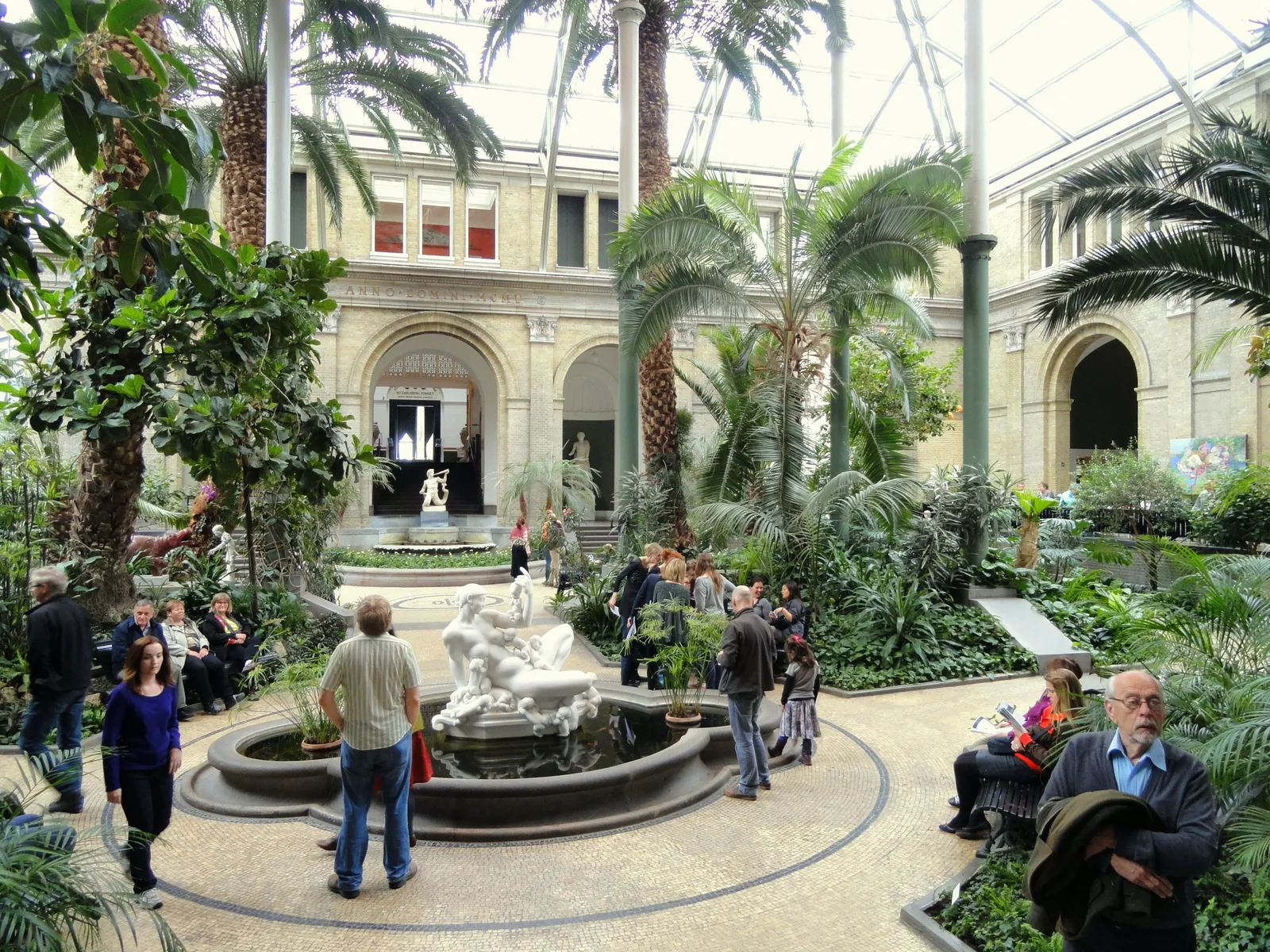 The Winter Garden inside the Ny Carlsberg Glyptotek in Copenhagen, tall palms and dense greenery around a stone fountain under a high glazed dome, with visitors sitting on benches along the edges