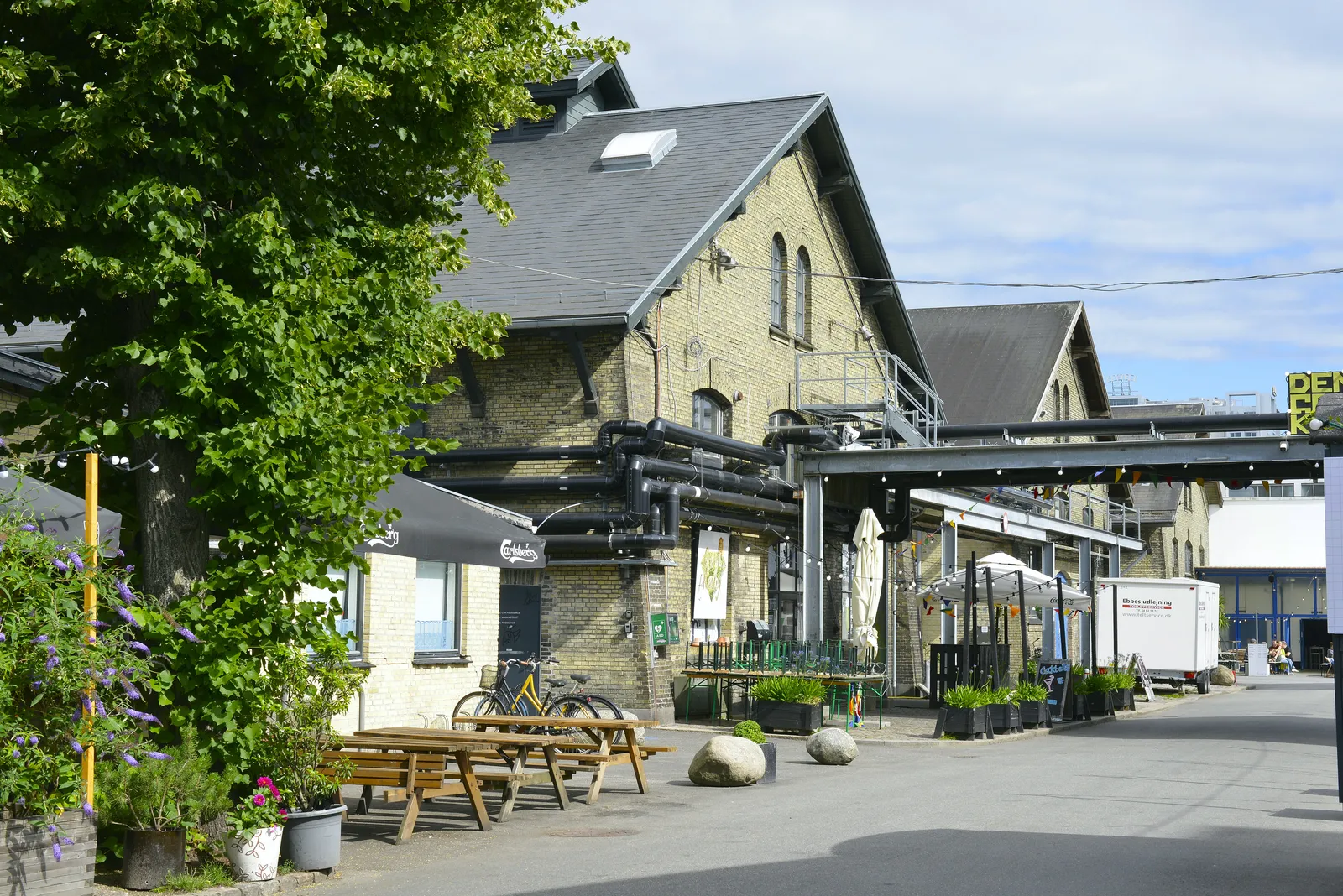A street through the old meatpacking district in Copenhagen, yellow brick sheds with steep slate roofs and exposed pipework, picnic tables and bicycles outside a cafe under a tree