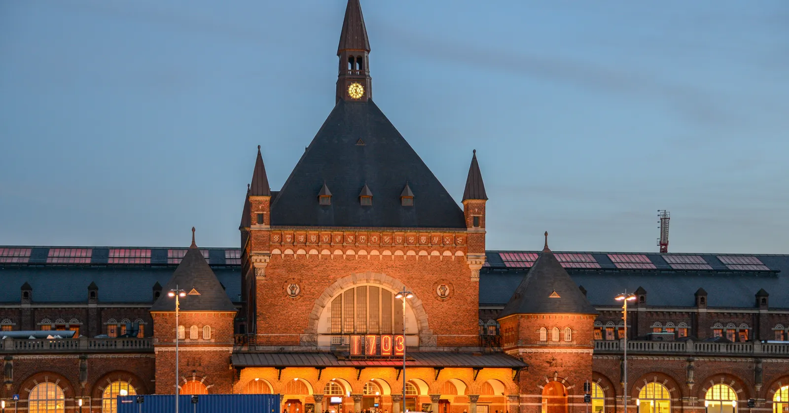 The front gable of Copenhagen Central Station at dusk, a steep dark roof over red brick with a gold-faced clock in the peak and a large arched window lit from inside below it