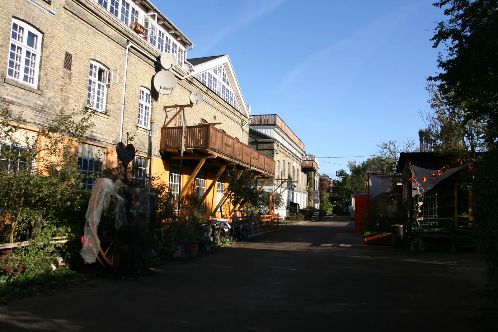 An unpaved lane inside Freetown Christiania in Copenhagen, a long yellow-brick former barracks block with an added timber balcony and staircase on one side, plants and painted sheds on the other