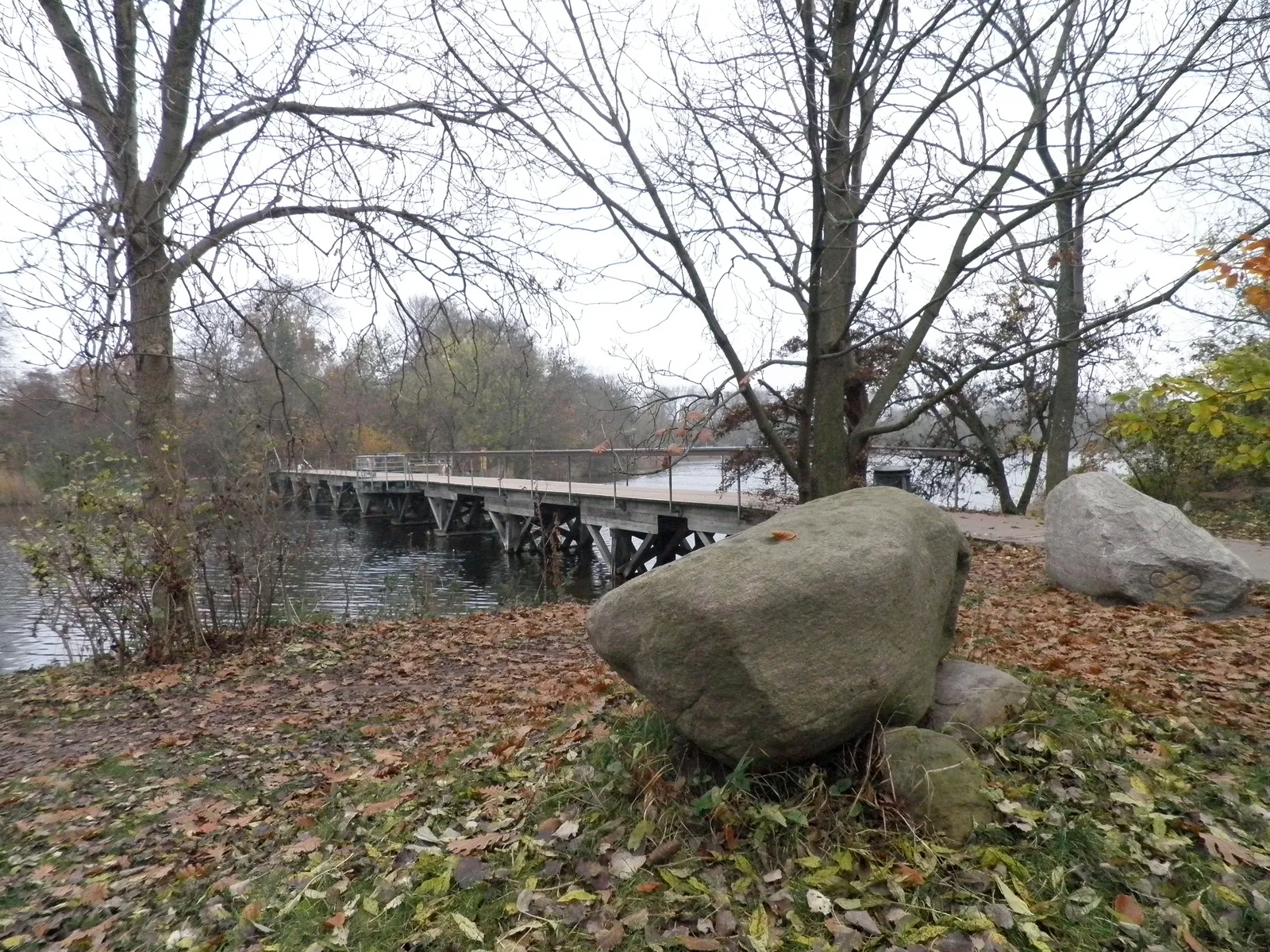 A narrow wooden footbridge crossing the still water of the old moat at Christianshavn in late autumn, bare trees on both banks, fallen leaves and large boulders on the near shore