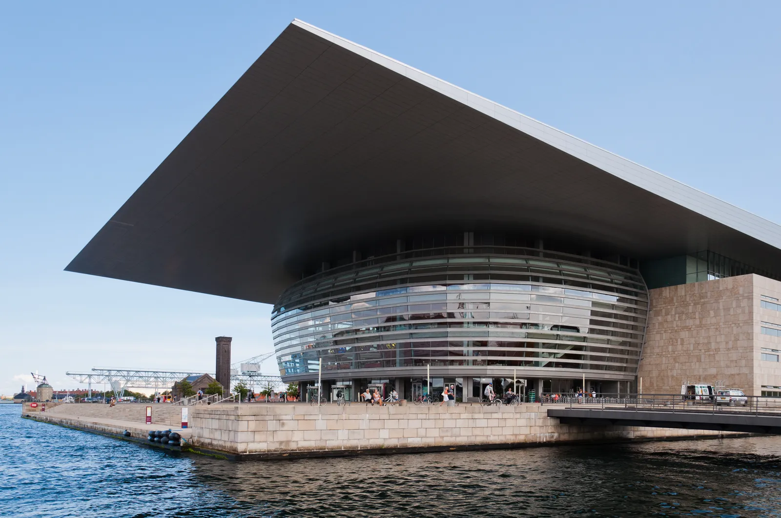 The Copenhagen Opera House on Holmen from across the water, a curved glass front under a flat roof that cantilevers far out over the quay, with the limestone base meeting the harbour