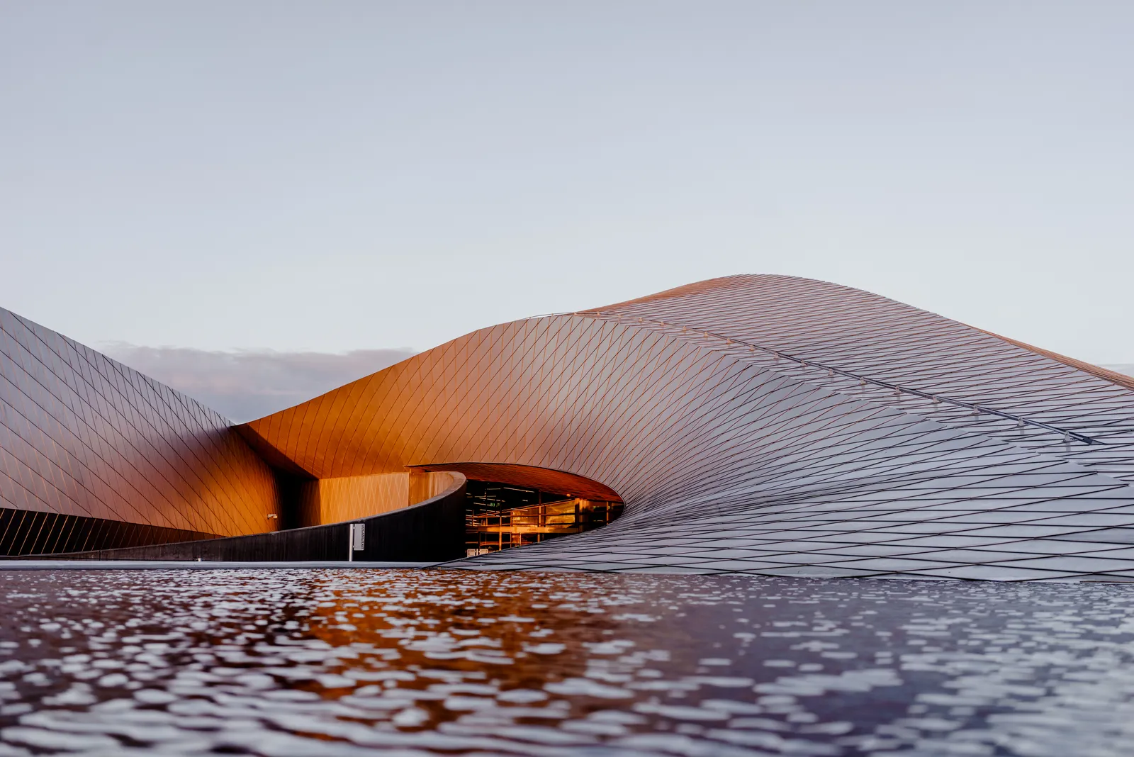 The National Aquarium of Denmark at Kastrup at dusk, a low building clad in overlapping metal shingles curving away in arms like the arms of a whirlpool, lit orange inside and reflected in the water in front of it