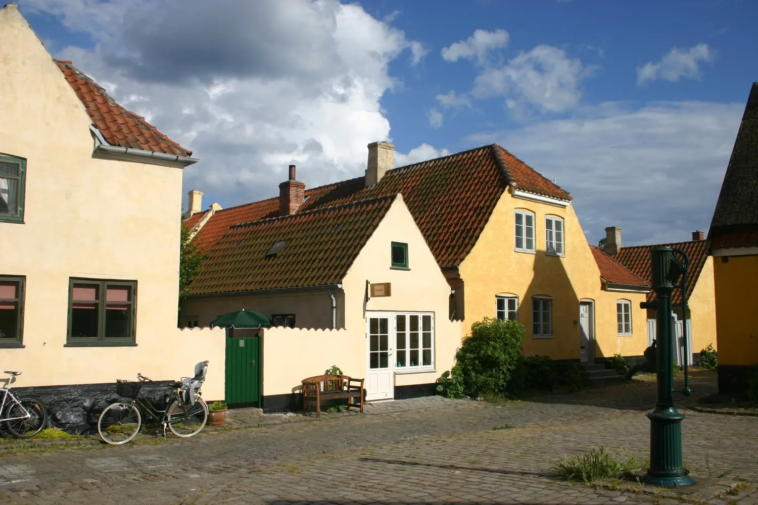A cobbled street corner in Dragør, low cream and yellow houses with red pantile roofs and dark green doors, bicycles leaning against a wall and a green cast-iron water pump in the foreground