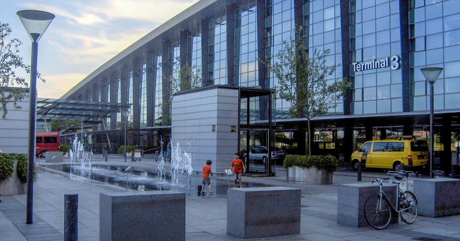 The entrance to Terminal 3 at Copenhagen Airport, a long glass front under a curved roof with the terminal number in blue lettering, a row of low fountains in the granite forecourt and a yellow taxi waiting at the kerb