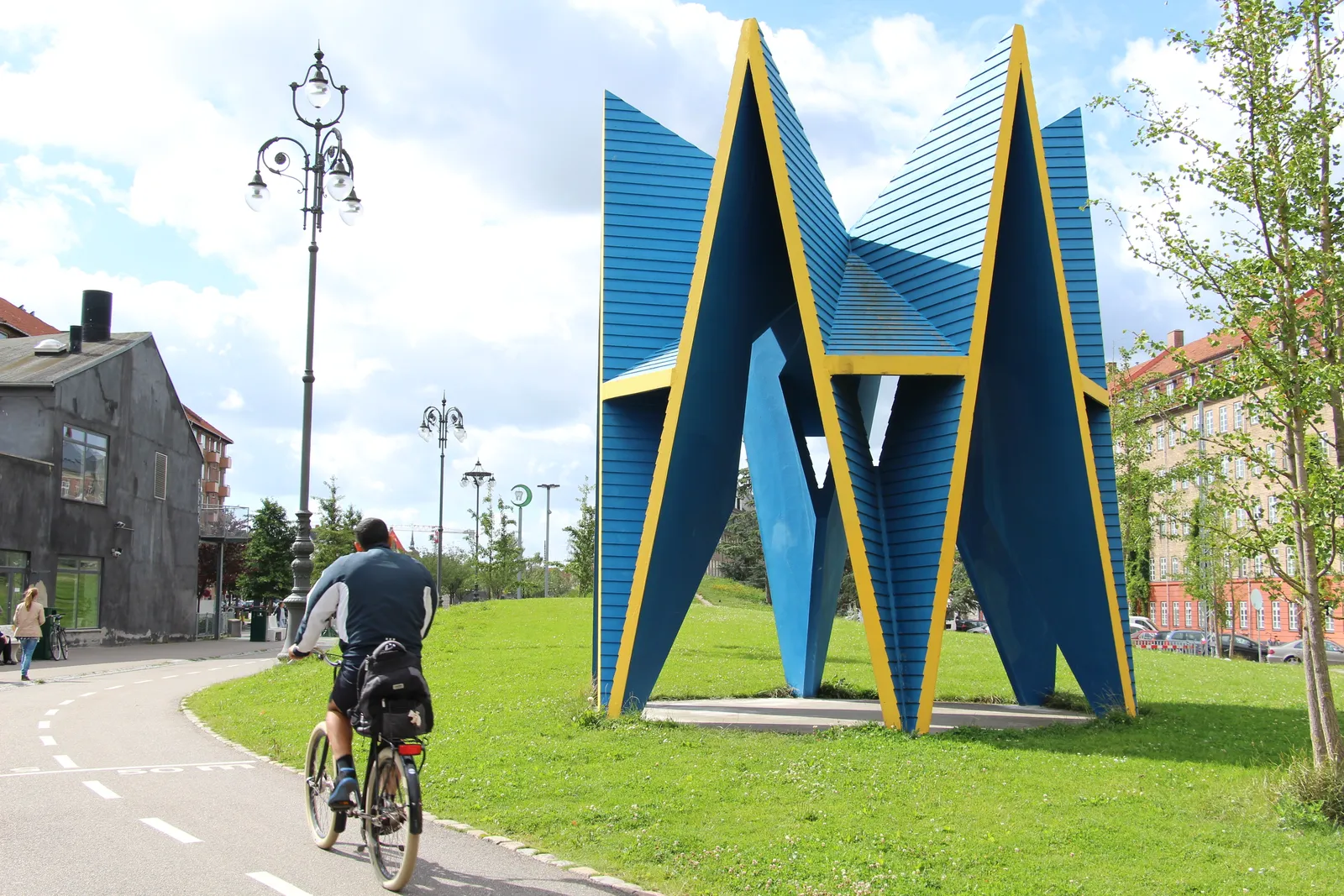 The green section of Superkilen park in Copenhagen, a tall blue and yellow steel pavilion standing on the grass beside a curving pink-surfaced cycle path, with a cyclist riding past and apartment blocks behind