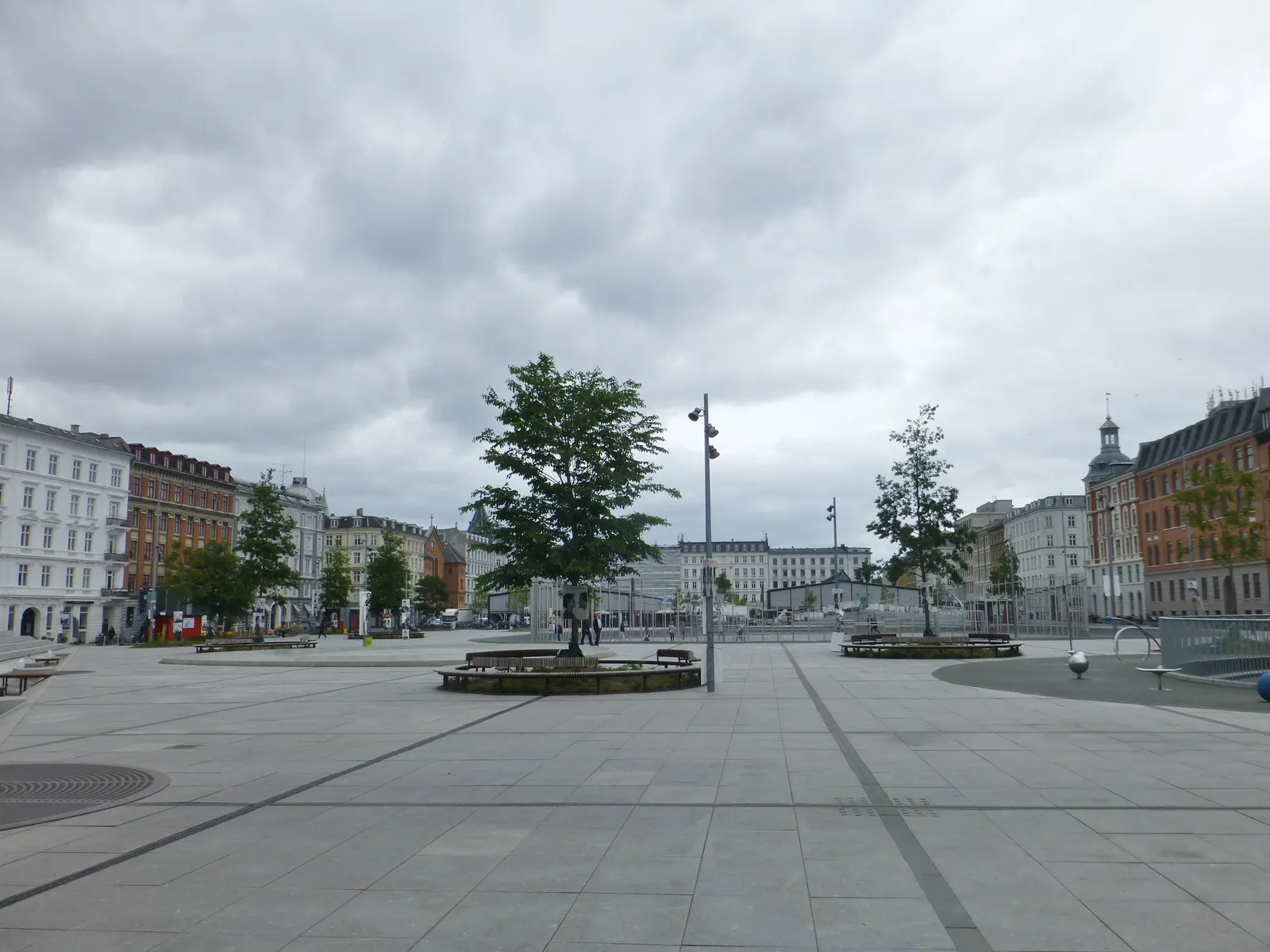 The open granite surface of Israels Plads in Copenhagen, pale paving running the width of the square with a raised circular planter and trees in the middle, and 19th-century apartment blocks along both sides