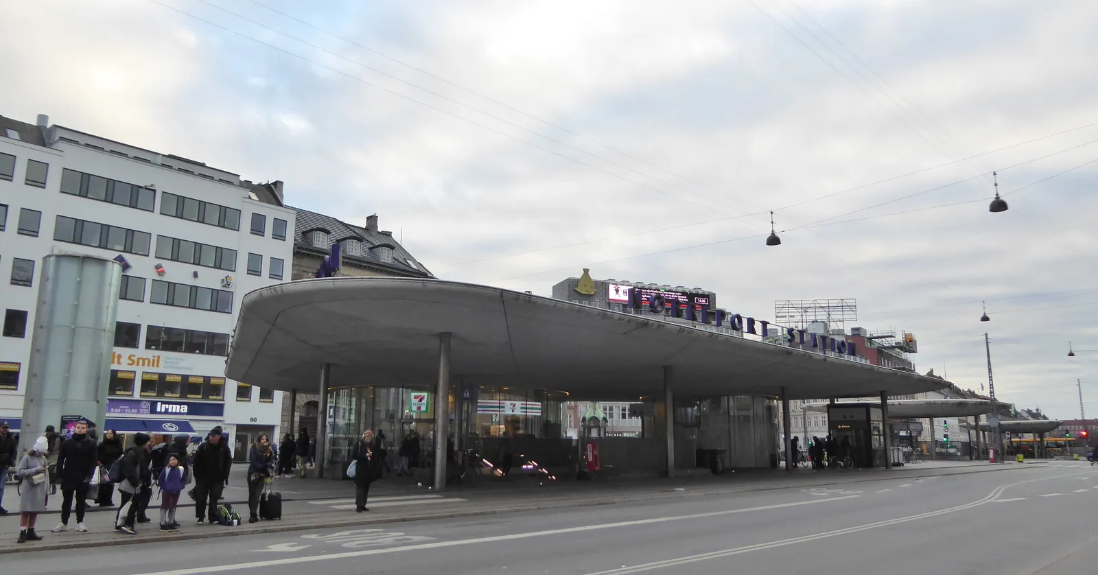 The street-level canopy at Nørreport station in Copenhagen, a long curved concrete roof on slim columns over the pavement, with waiting passengers underneath and the station name spelled out along its edge