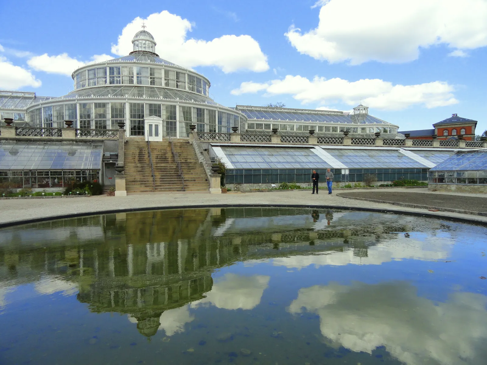 The Palm House in Copenhagen's Botanical Garden, a domed Victorian glasshouse on a cast-iron frame with lower wings either side, reflected in the still pond in front of it under a blue sky