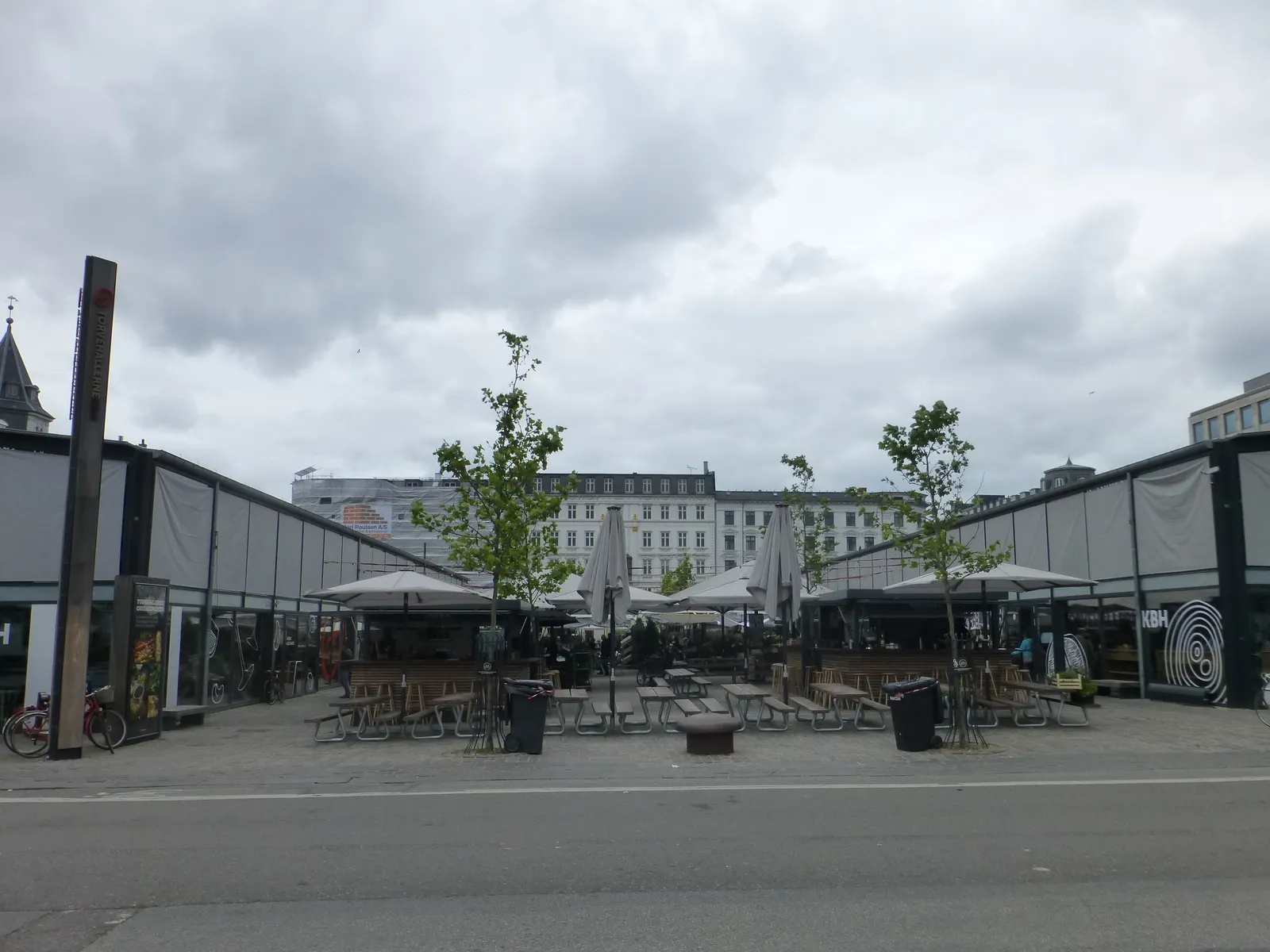 The two market halls of Torvehallerne in Copenhagen facing each other across an open paved strip, dark steel frames filled with glass, with cafe tables, parasols and young trees set out between them