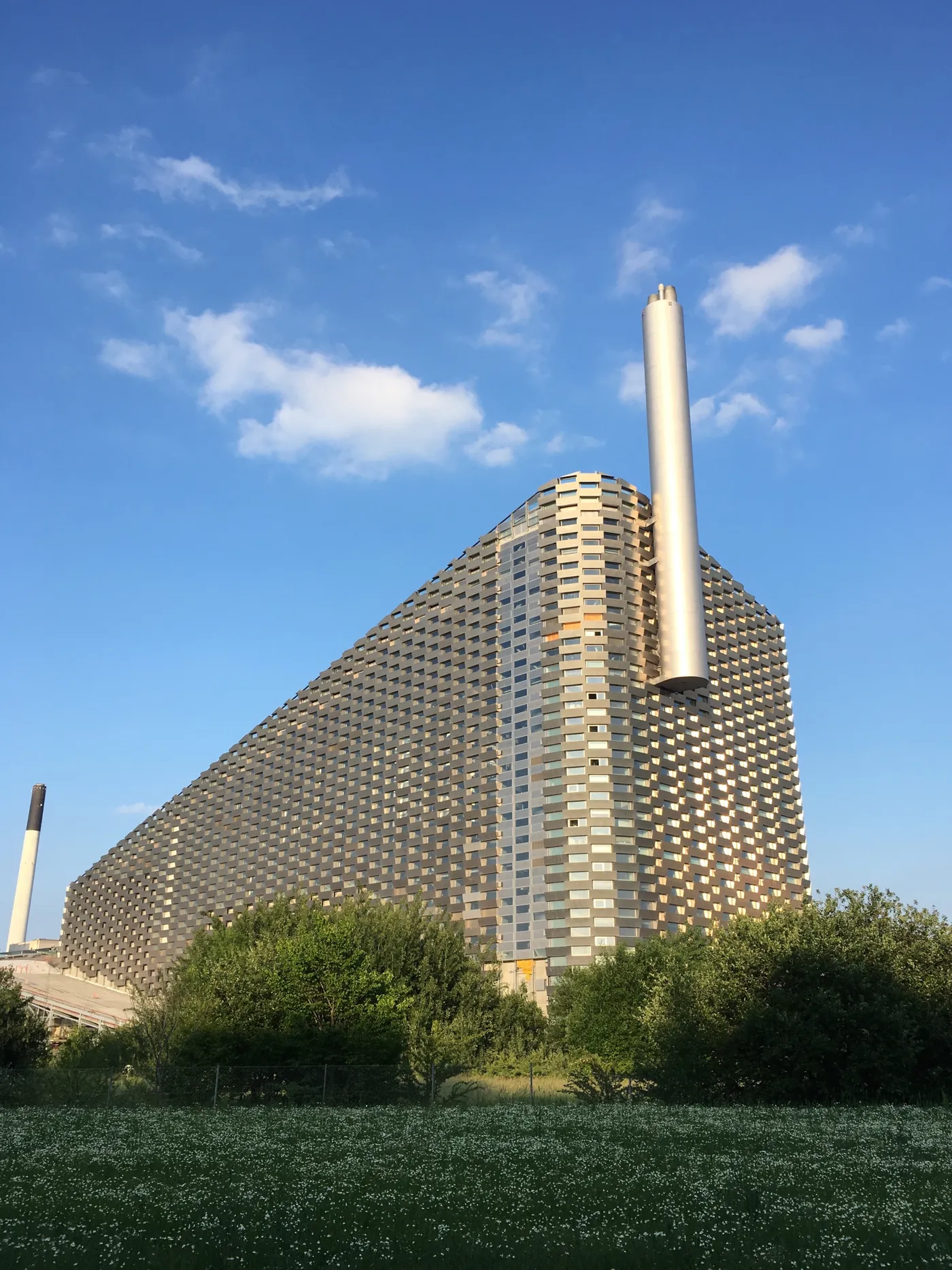 The Amager Bakke waste-to-energy plant in Copenhagen, a huge block clad in stacked aluminium panels sloping steeply from ground level to a flat top, with a wide white chimney rising beside it against a blue sky