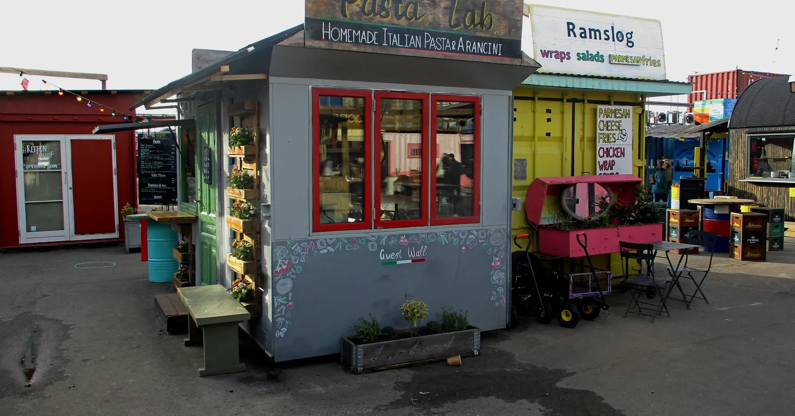 A food stall at the Reffen street food market in Copenhagen, a small painted hut with a serving window and menu boards, stacked shipping containers and further stalls behind it on open shipyard concrete