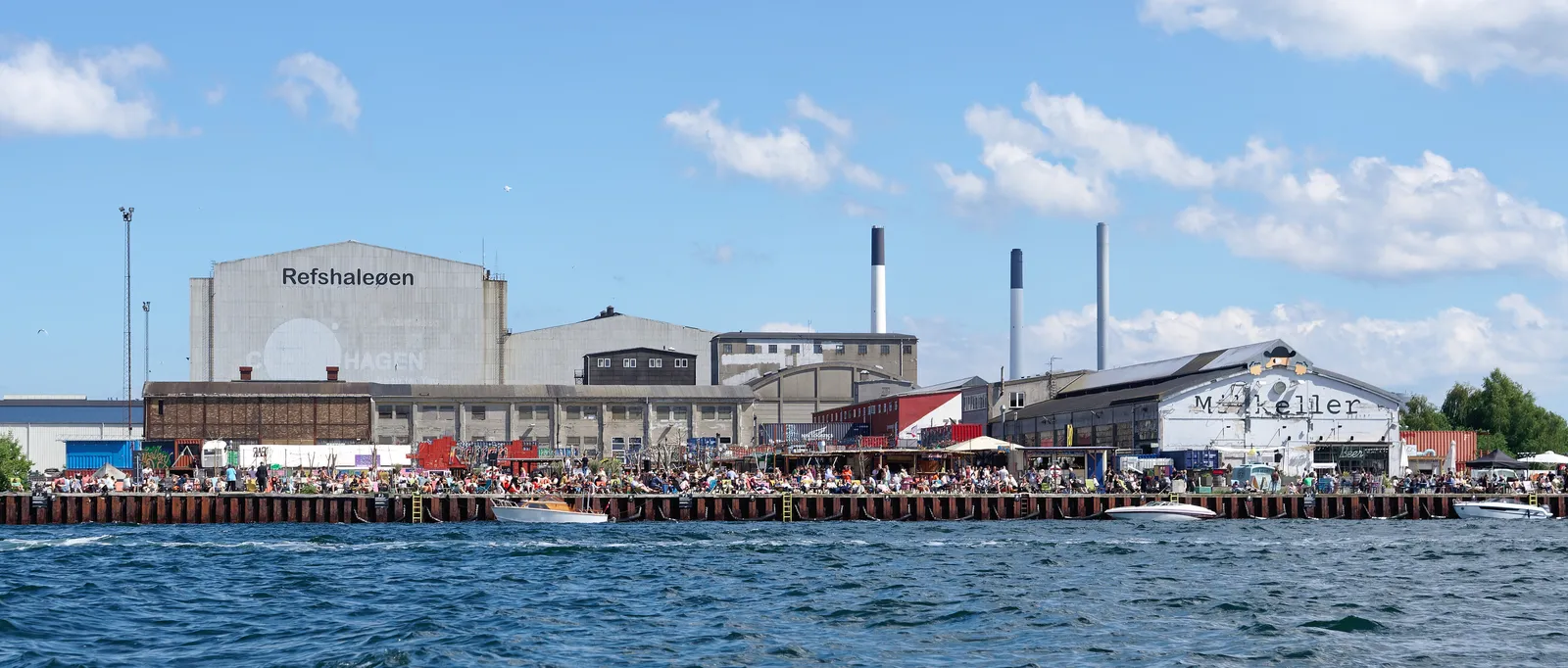 The industrial waterfront of Refshaleøen in Copenhagen seen from the harbour, long shipyard sheds with the island's name painted across one gable, chimneys behind, and people lining the quay in the sun